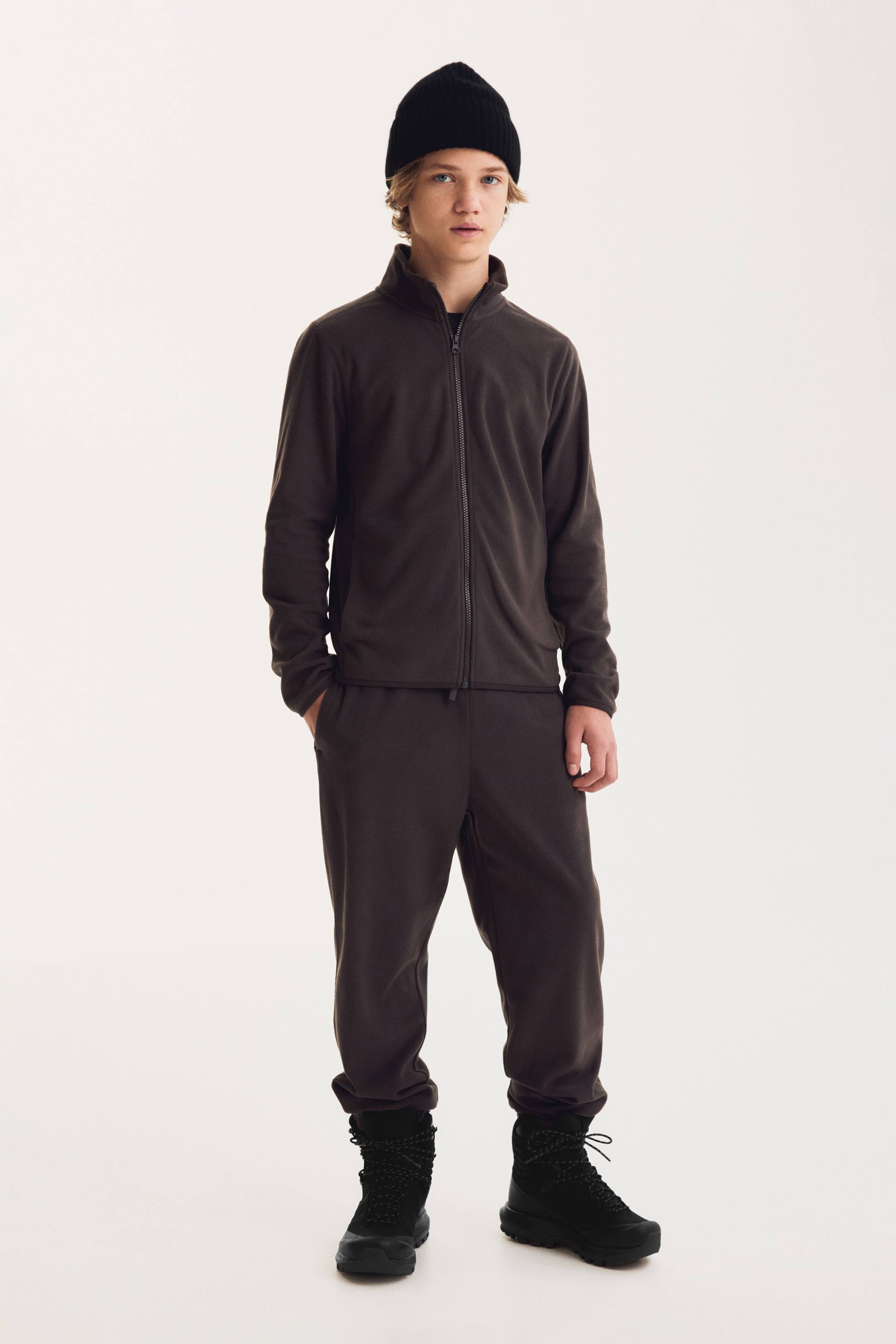 Ver imagen más grande: Se muestran unos pantalones deportivos de forro polar en marrón oscuro vistos de frente, con un corte regular y bajos elásticos fruncidos por encima de unas botas negras. Los pantalones se coordinan con una chaqueta a juego con cremallera, destacando su textura suave y ligera y su caída relajada.