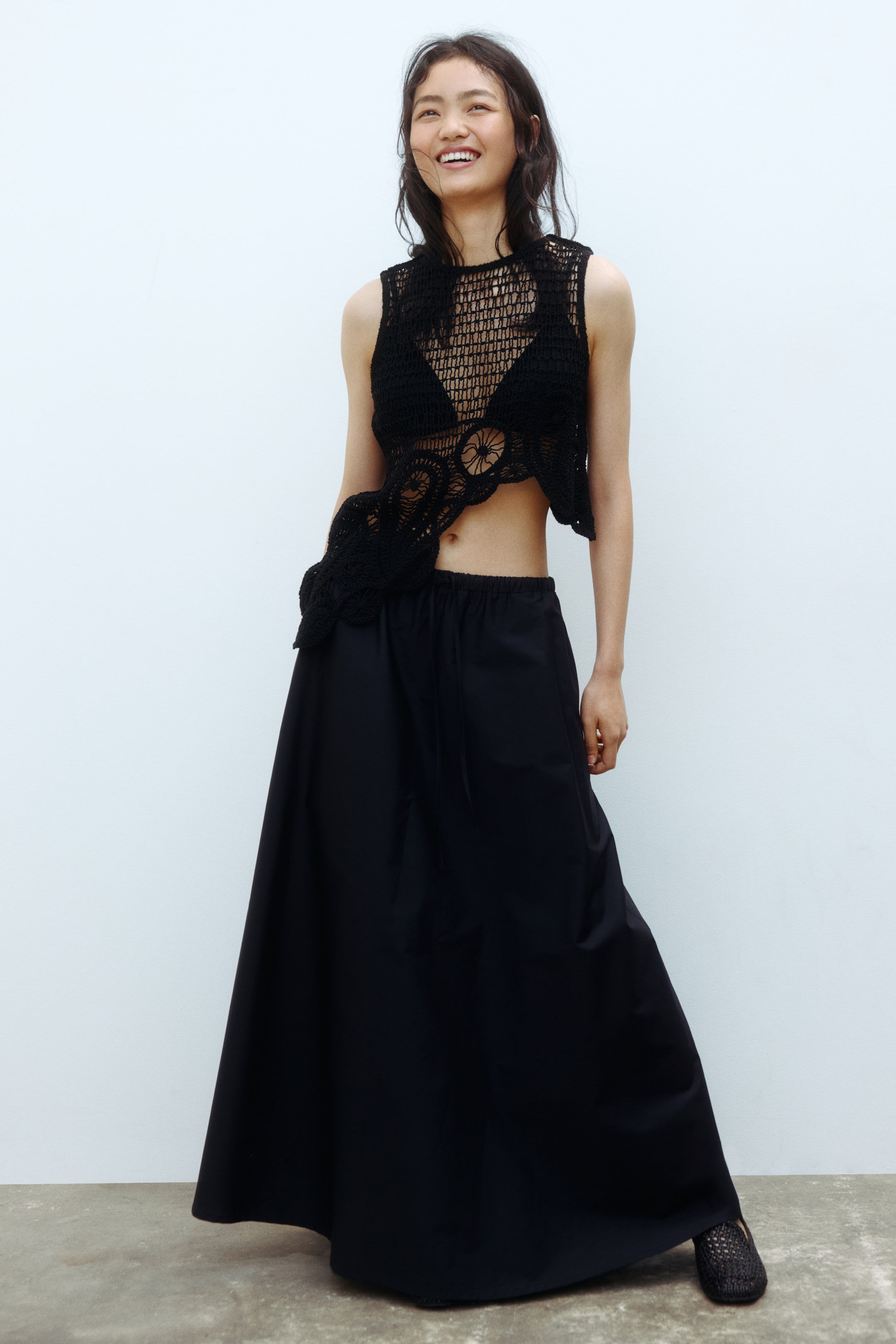 Voir une image plus grande: Un haut court sans manches en crochet noir est assorti à une jupe longue fluide noire, tous deux vus de face. Le haut présente un tricot ouvert avec des motifs circulaires et un ourlet asymétrique, porté sur un soutien-gorge noir visible. La jupe est dotée d'une taille élastiquée avec cordon de serrage et d'une silhouette évasée en ligne A.