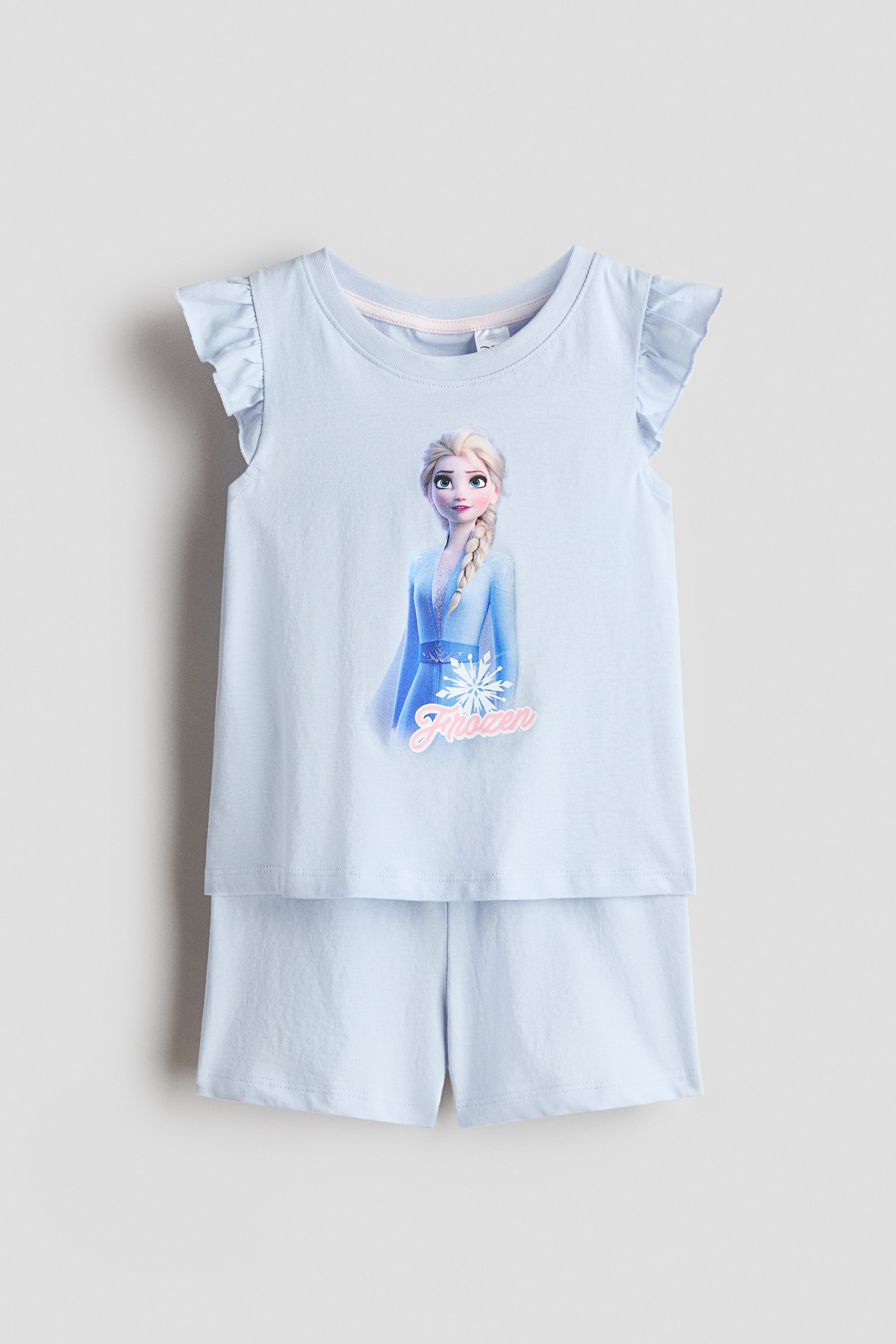 Vis større bilde: Et lyseblått jersey-pyjamasett presenteres forfra, bestående av en topp med rund halsutringning og flagrende ermer, samt matchende shorts. Toppen viser et grafisk trykk av Elsa, ordet "Frozen", og en snøfnugg.