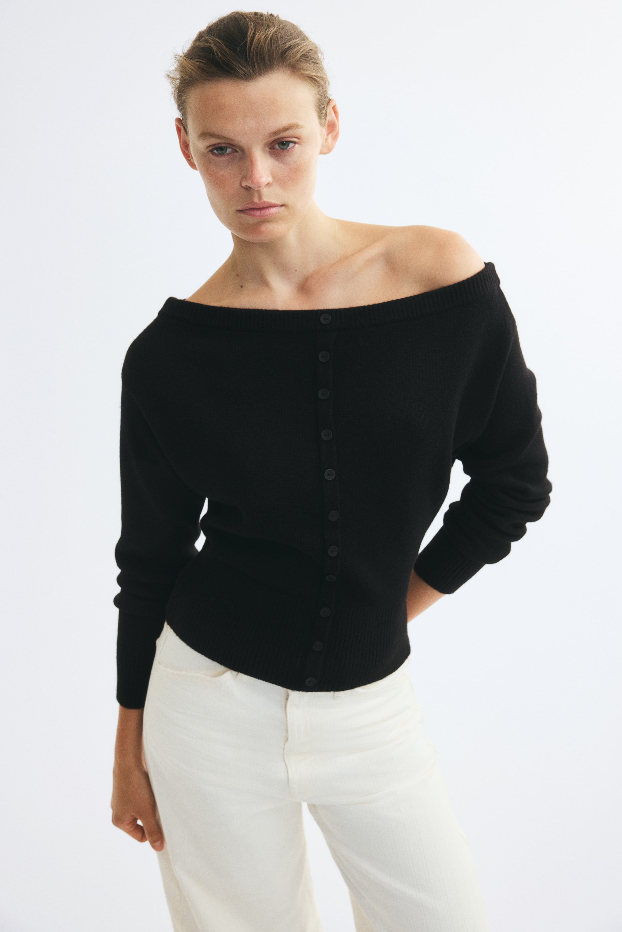 View larger image: A black knit cardigan featuring a button-down front, ribbed trim, and long sleeves is being worn by the model with a wide neckline draped off one shoulder. The cardigan is paired with off-white trousers.