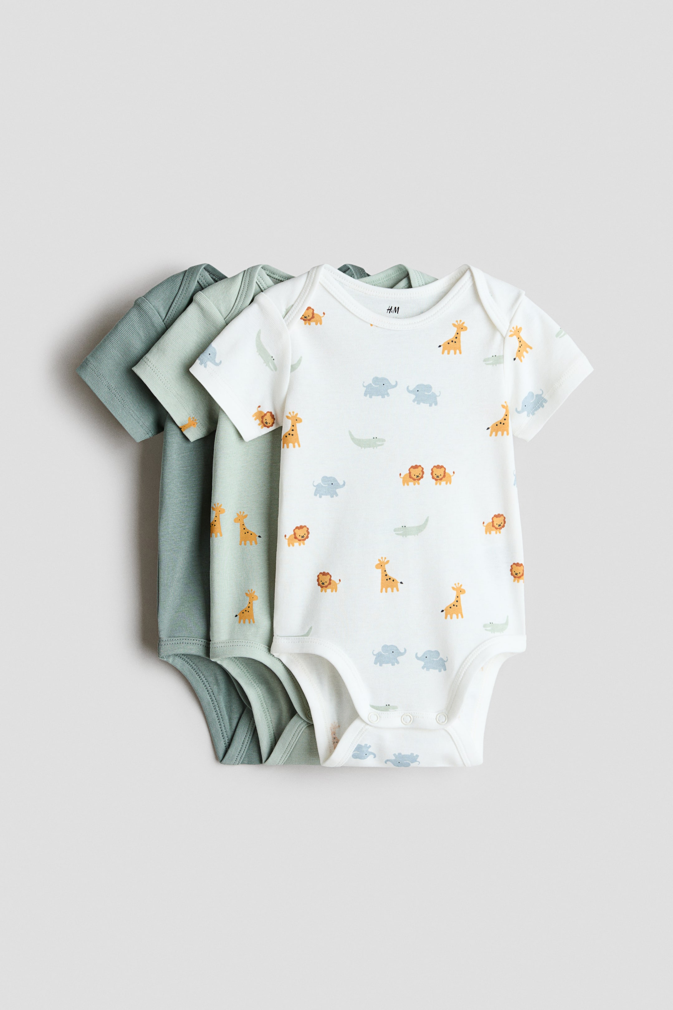 View larger image: Short-sleeved bodysuits in soft cotton jersey, featuring an envelope neck and press-studs at the bottom, are showcasing their front sides. The front bodysuit is white with an all-over print of cartoon giraffes, lions, elephants, and crocodiles, while two solid green bodysuits are layered behind it.