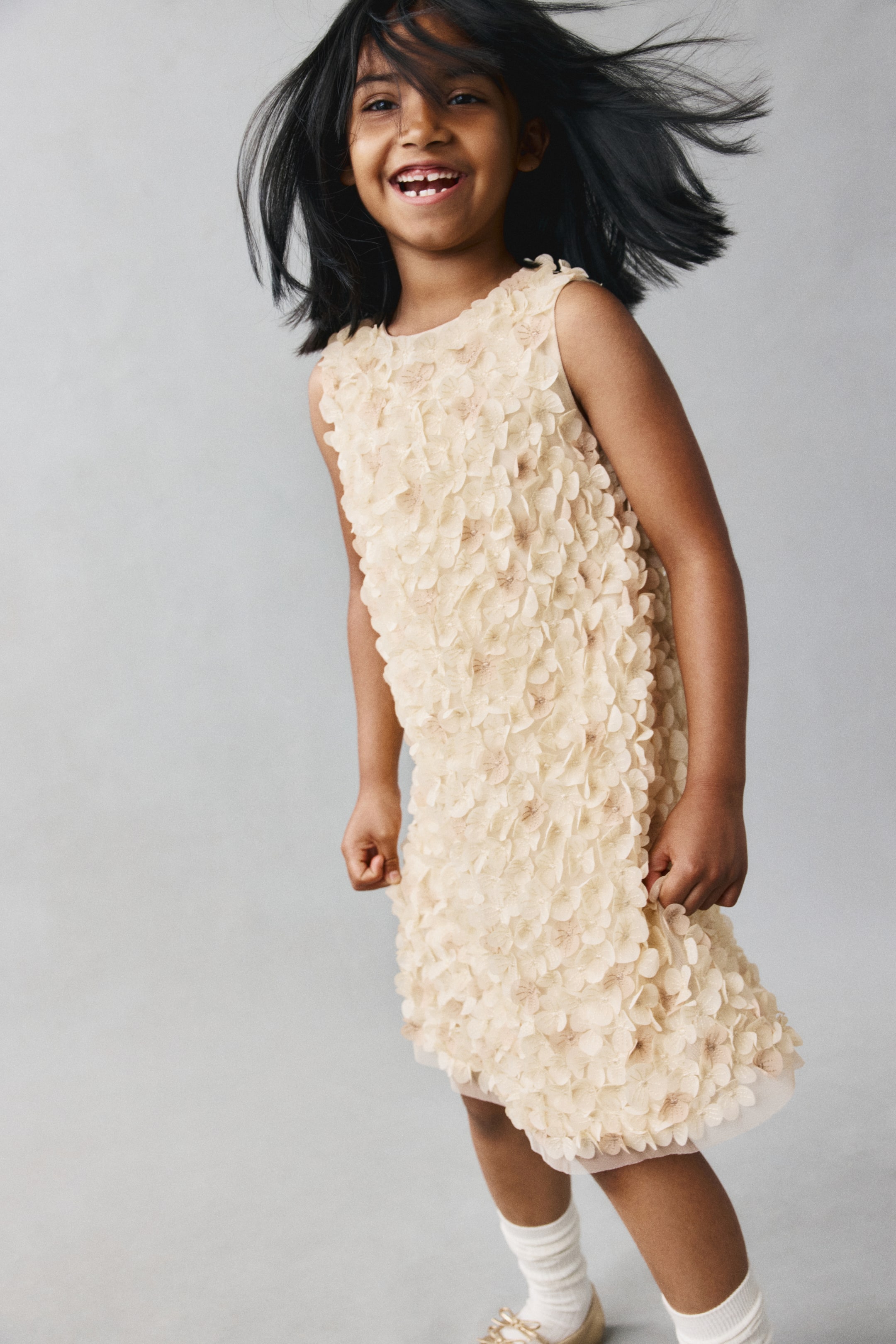 View larger image: A sleeveless cream-coloured shift dress is shown from the front, featuring a round neckline and an all-over texture of layered 3D floral appliqués. The dress has a straight silhouette with a sheer underlayer peeking out slightly at the hem.