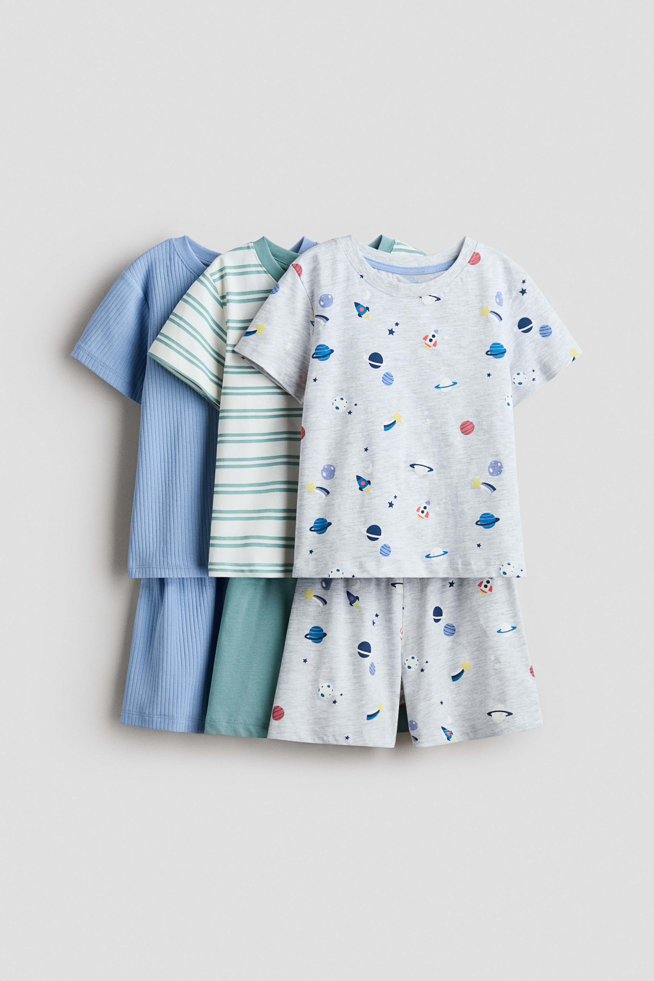 View larger image: Three sets of pyjamas are layered, each featuring a T-shirt and matching shorts. The front set includes a light grey T-shirt with a space-themed print, short sleeves, and a ribbed neckline, paired with matching shorts. Behind it, a white T-shirt with green horizontal stripes and matching shorts are visible, followed by a light blue ribbed T-shirt and ribbed shorts.