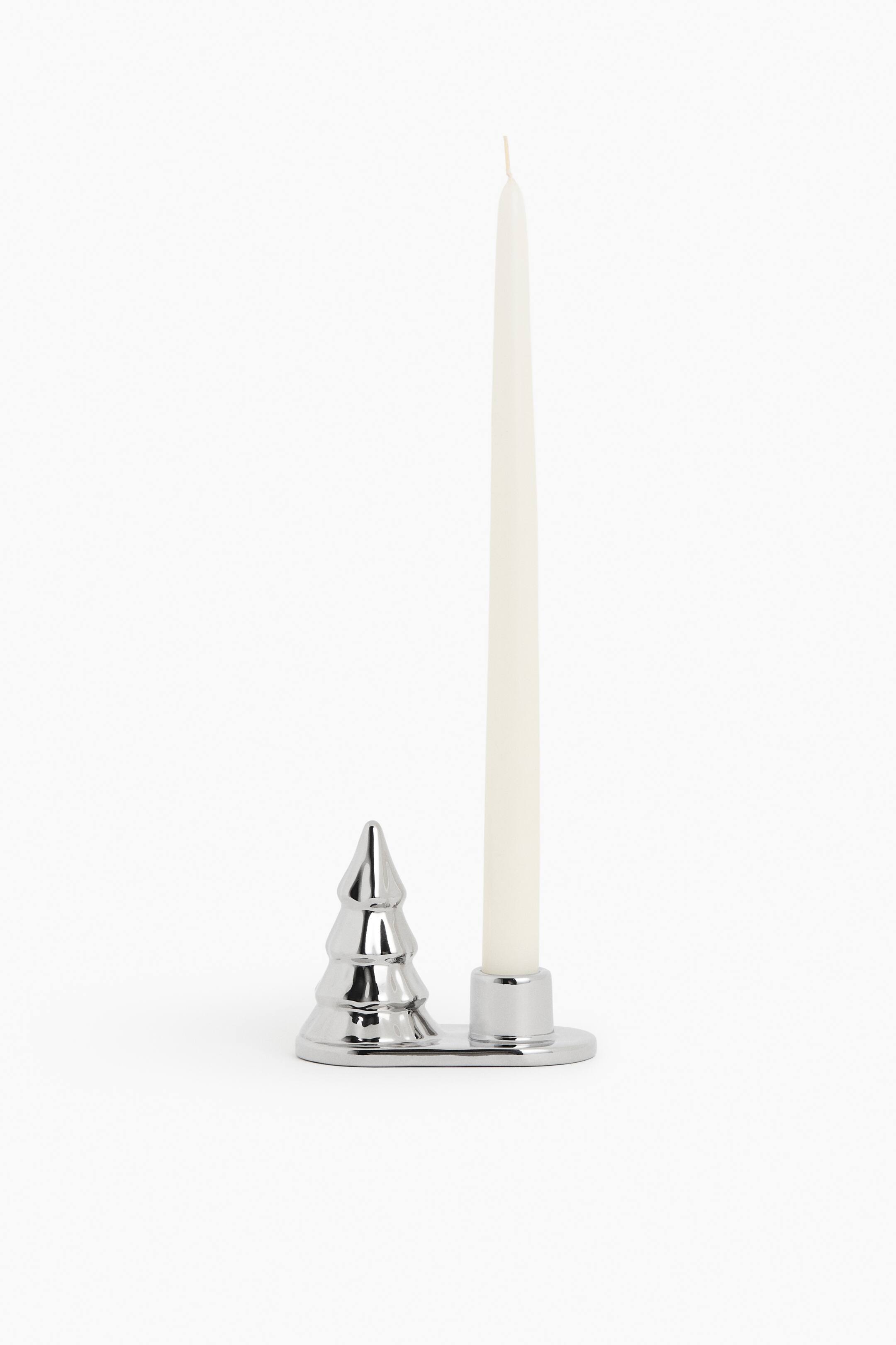 View larger image: A silver-toned candle holder features a polished finish and a flat base, with a decorative Christmas tree shape and a single candle well. A white taper candle stands upright in the well, with the holder presented at a slight angle.