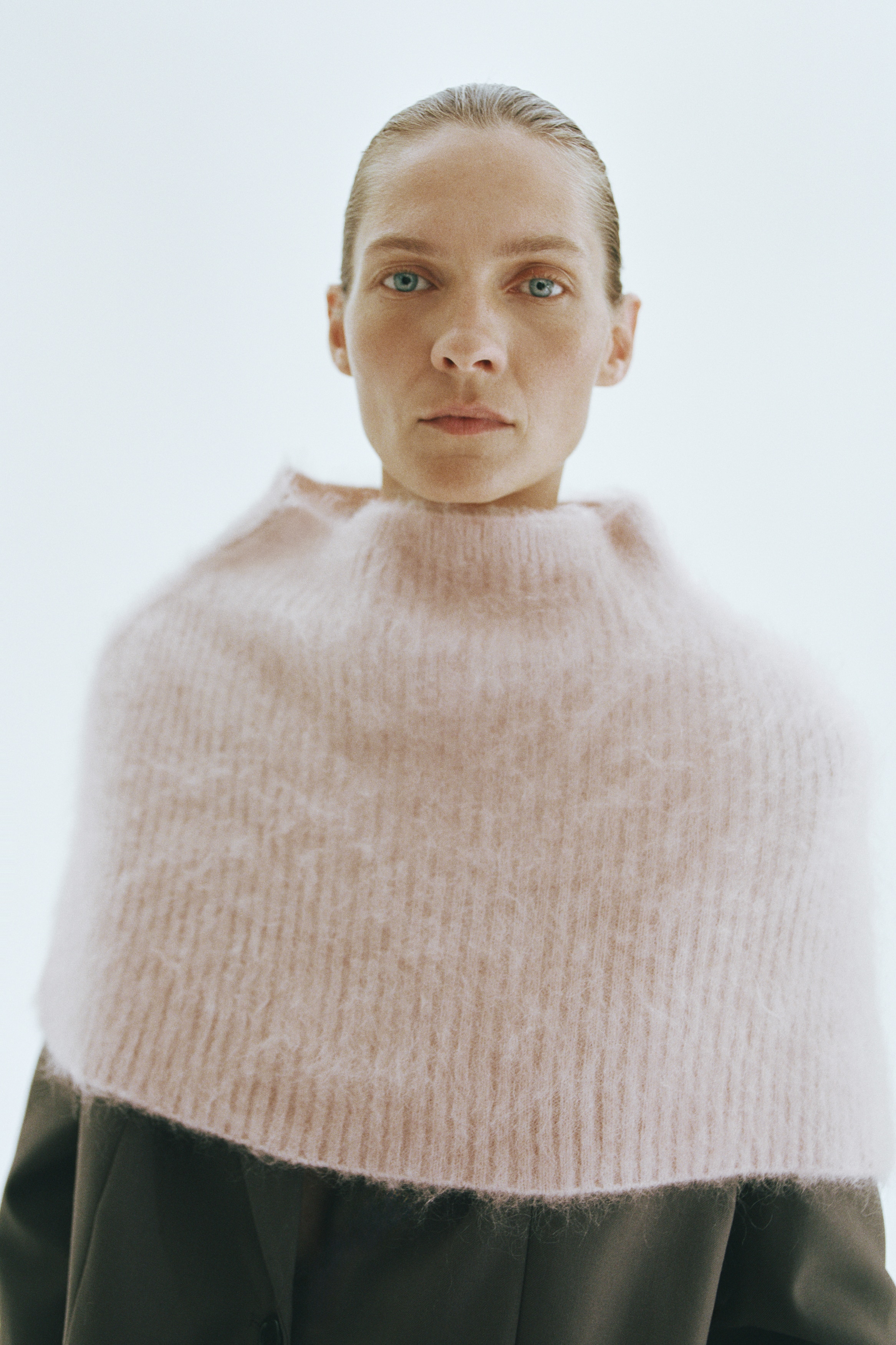 A light pink mohair-blend rib-knit cape is shown from the front, featuring a mock turtleneck and a flared silhouette that drapes over the shoulders. The garment displays a soft, fuzzy texture with vertical ribbed detailing throughout.