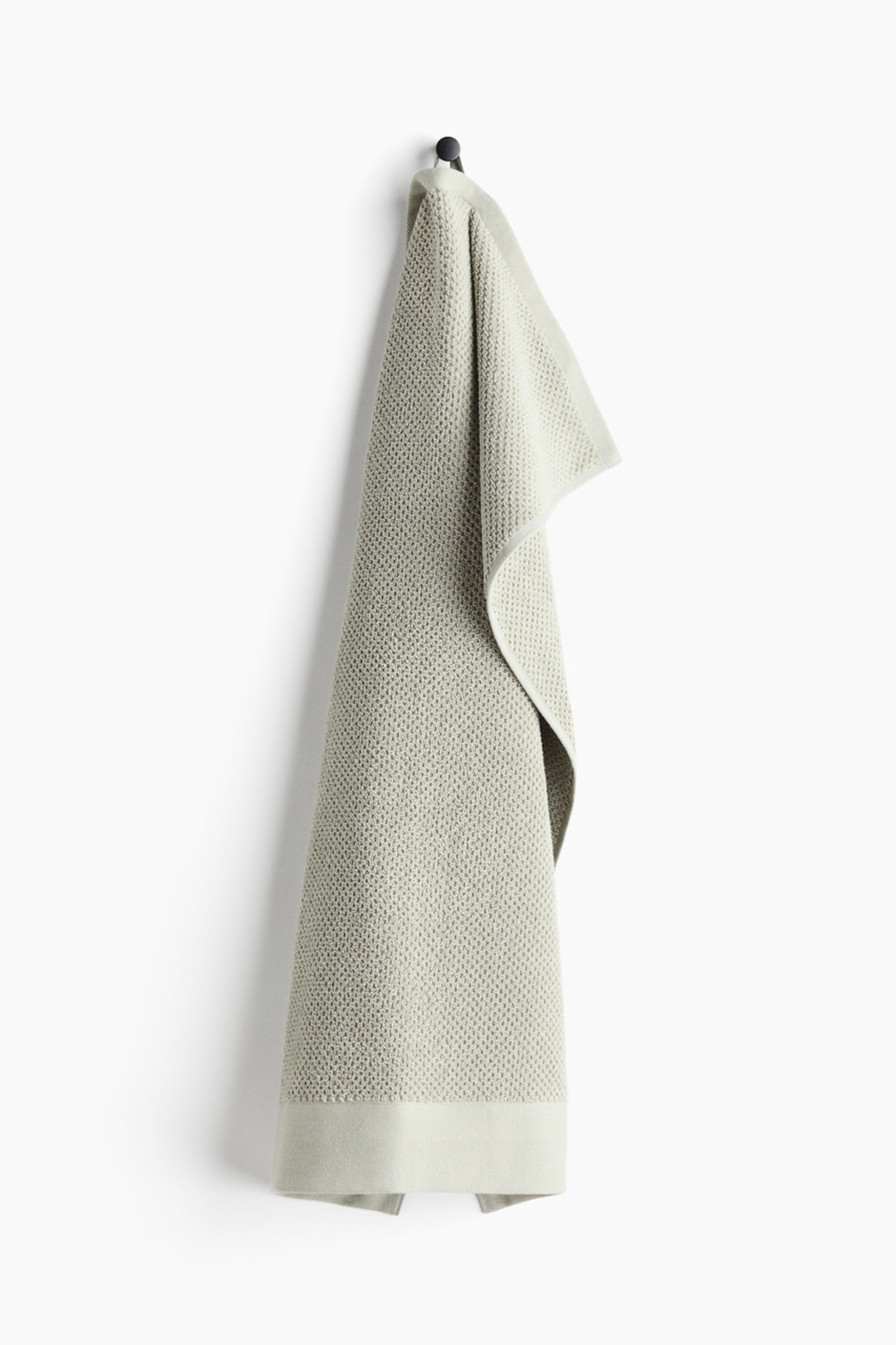 View larger image: A light green hand towel with a waffle weave texture is hanging vertically, folded over a black hook. A smooth, flat band is visible along the bottom edge, and a hanging loop on its short side is securing it at the top.