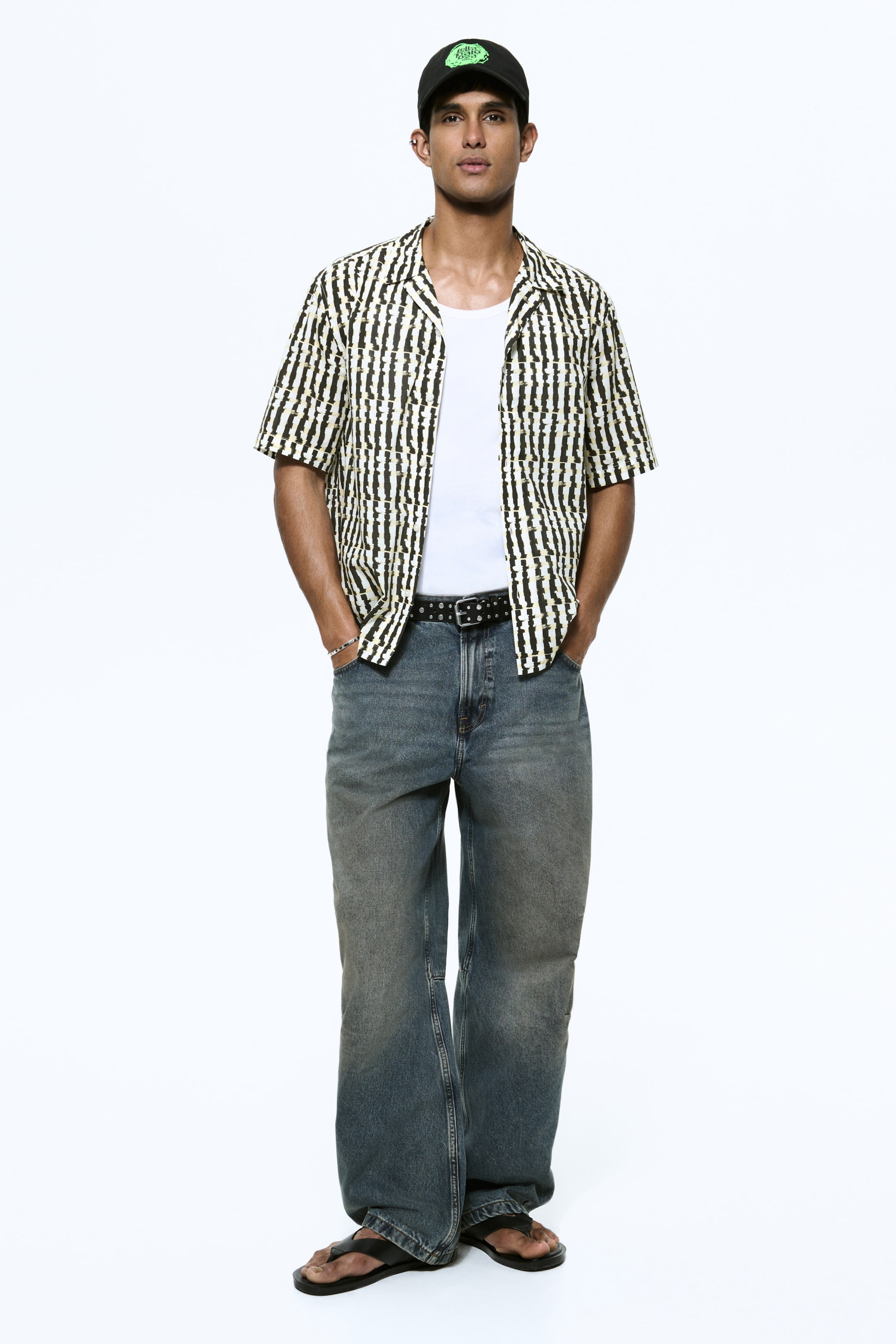 View larger image: A black baseball cap with a green graphic is worn with a short-sleeved shirt with an open collar and irregular black-and-yellow vertical stripes on a white base, layered over a white crew-neck T-shirt. Loose-fitting blue denim jeans with a faded wash are cinched at the waist by a black studded belt, and black flip-flops are worn.