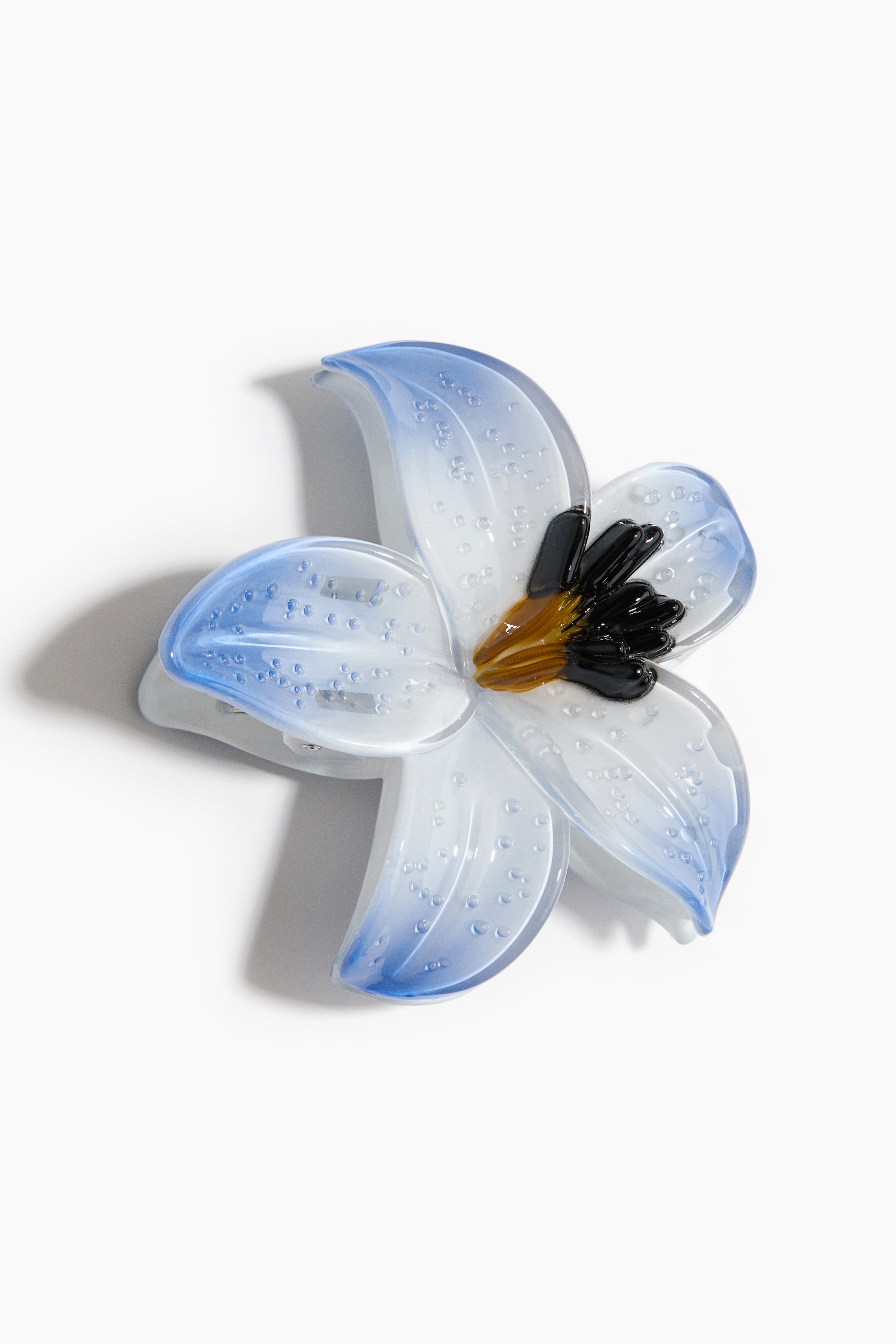 Voir une image plus grande: Une pince à cheveux en forme de fleur est orientée légèrement vers le haut et à droite, mettant en valeur ses pétales translucides blancs avec des pointes bleues. De petites textures en forme de bulles sont visibles sur les pétales brillants, et le centre présente des étamines de couleur noire et ambre.