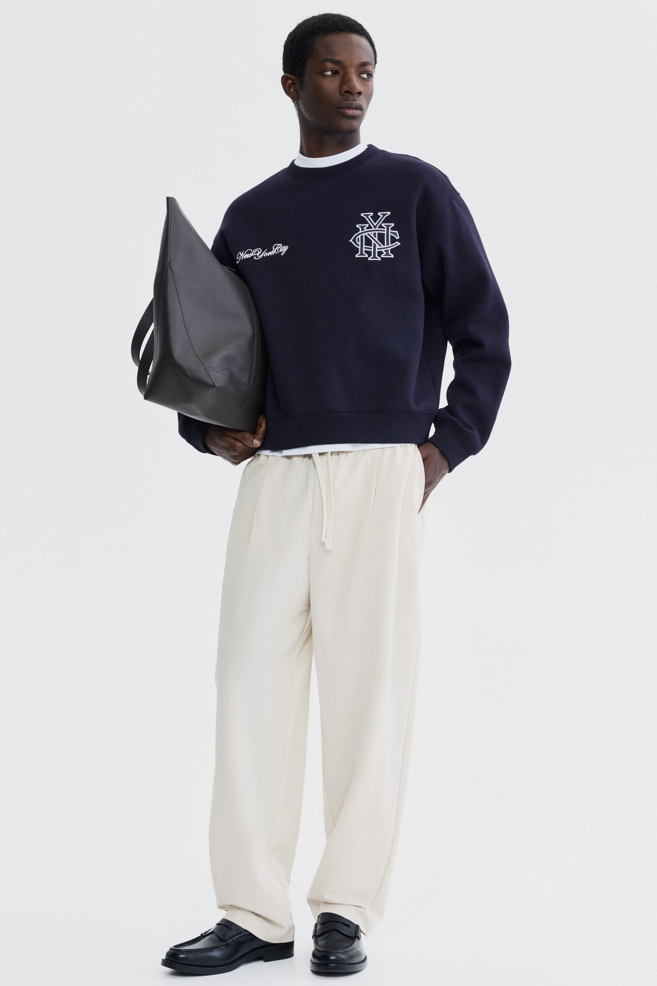 Ver imagem maior: Uma sweatshirt azul-marinho com gola redonda, com o texto "New York City" bordado a branco e um monograma entrelaçado na frente, está a ser usada sobre uma t-shirt branca e combinada com calças largas de cintura com cordão em creme. Mocassins pretos são visíveis nos pés, e uma mala tote preta angular está a ser segurada na mão esquerda.