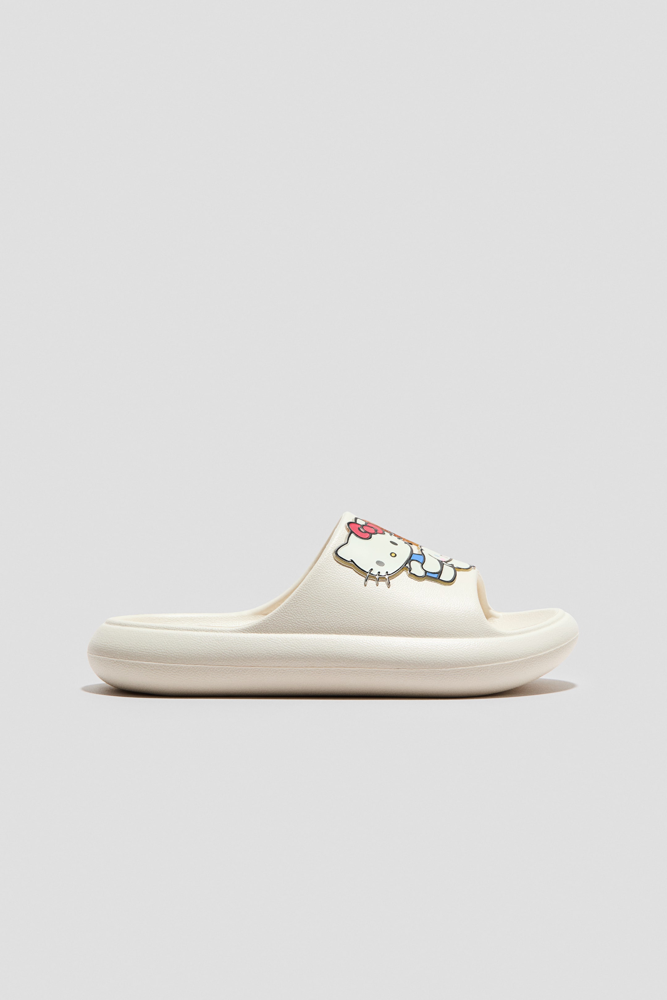 Agrandir l'image: Une chaussure de piscine moulée couleur crème est présentée en vue latérale, avec une large bride ornée d'un motif imprimé. Le motif représente un personnage félin blanc avec un nœud rose et une salopette bleue, semblant faire face vers l'avant.