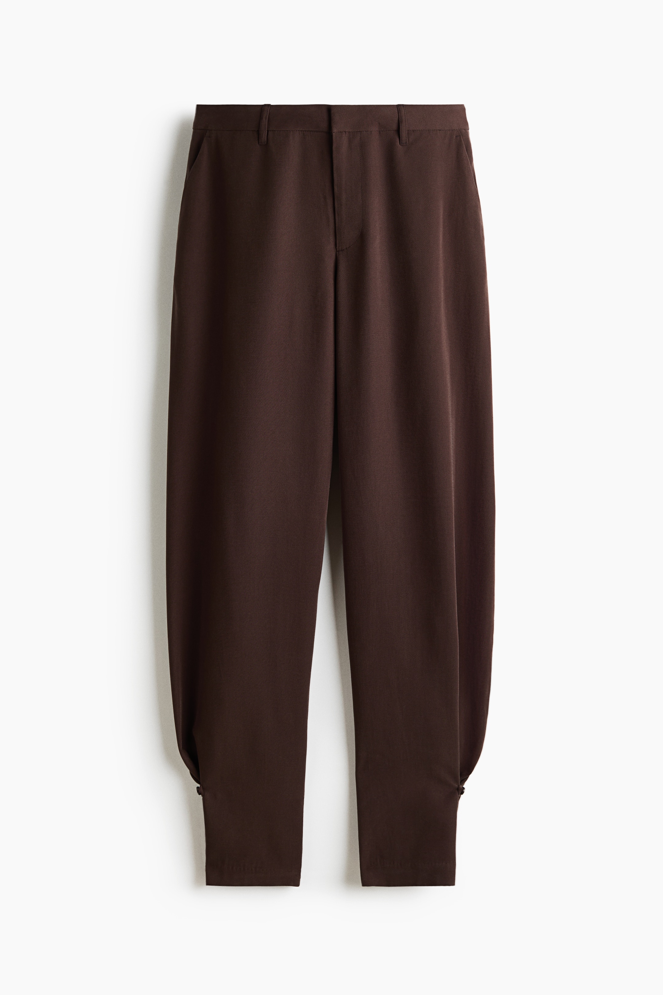 Visualizza immagine più grande: Questi pantaloni sartoriali marrone scuro sono mostrati frontalmente, con vita regolare con passanti per cintura e tasche laterali oblique. Il design presenta una silhouette a gamba barrel che si restringe alle caviglie, con un piccolo dettaglio con bottone e asola che crea un effetto arricciato.