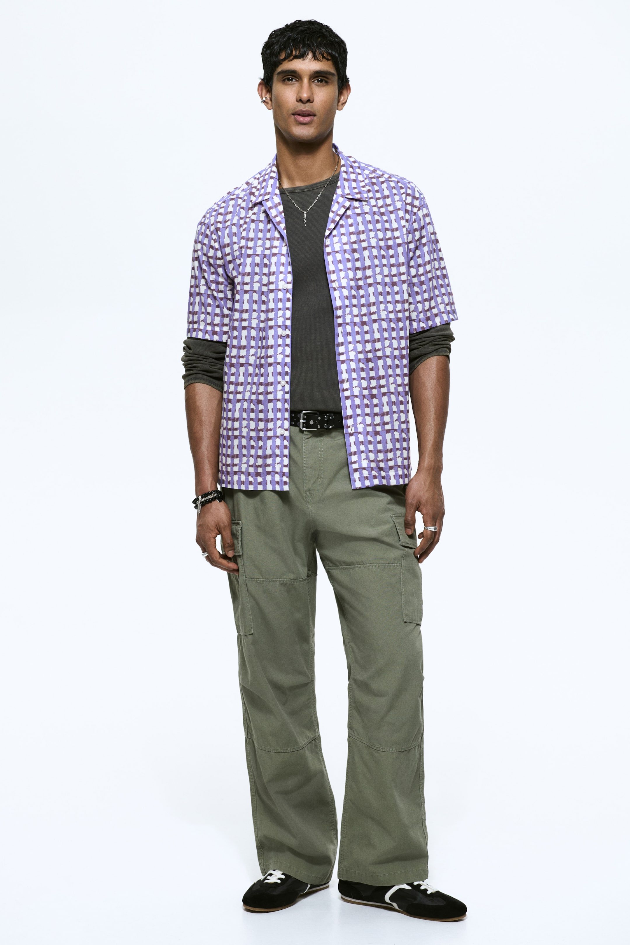 View larger image: Olive green cargo trousers are shown from the front, with a relaxed, straight-leg fit. They feature large flap cargo pockets on the sides, visible knee seams, and a black belt threaded through the belt loops, with the trousers falling to the ankles.