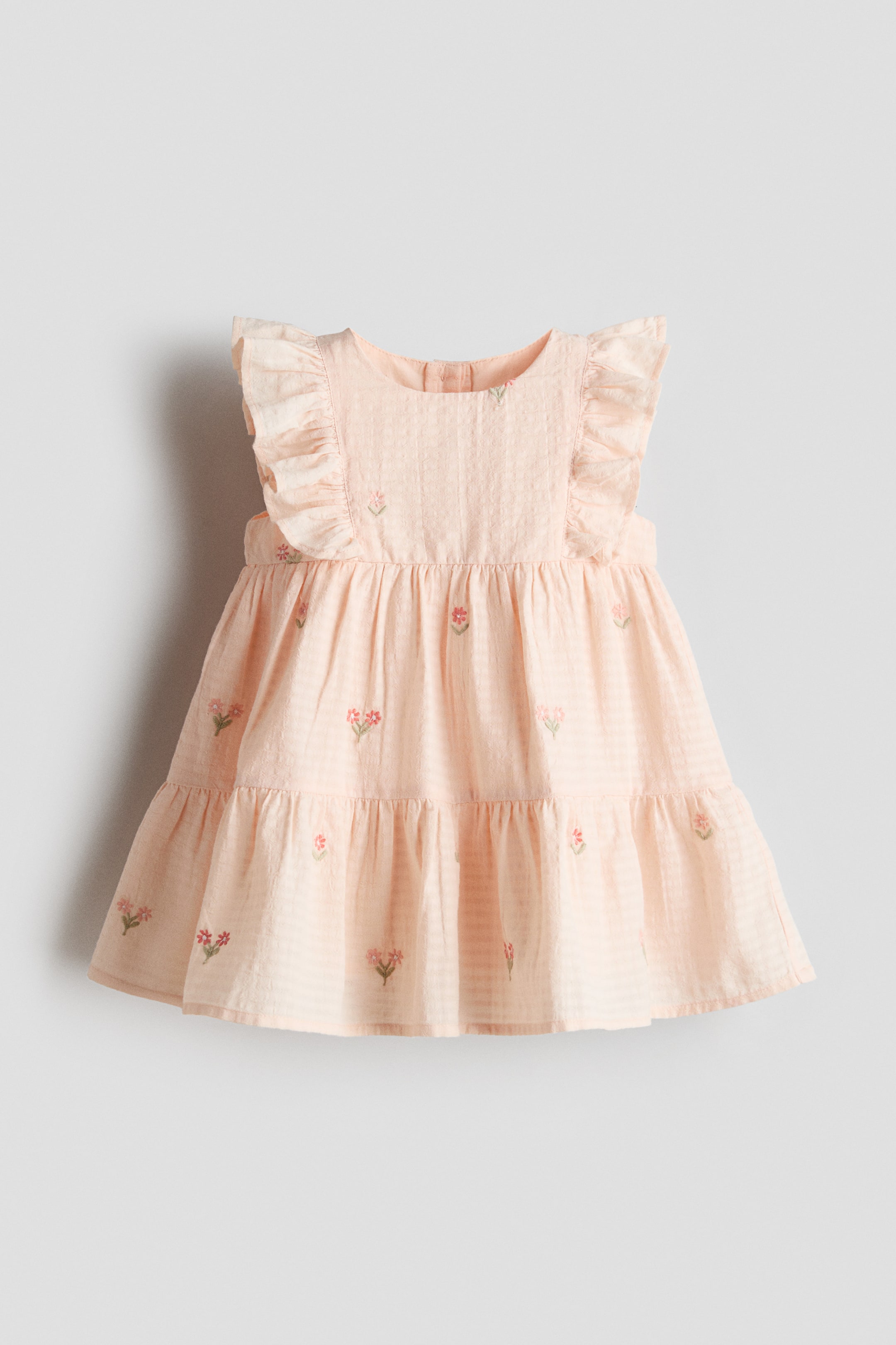 View larger image: A light peach baby dress showcases a gentle horizontal striped texture, featuring a rounded neckline, ruffled cap sleeves, and a tiered skirt. Delicate pink and green floral embroidery is scattered throughout the entire dress, presented from the front.