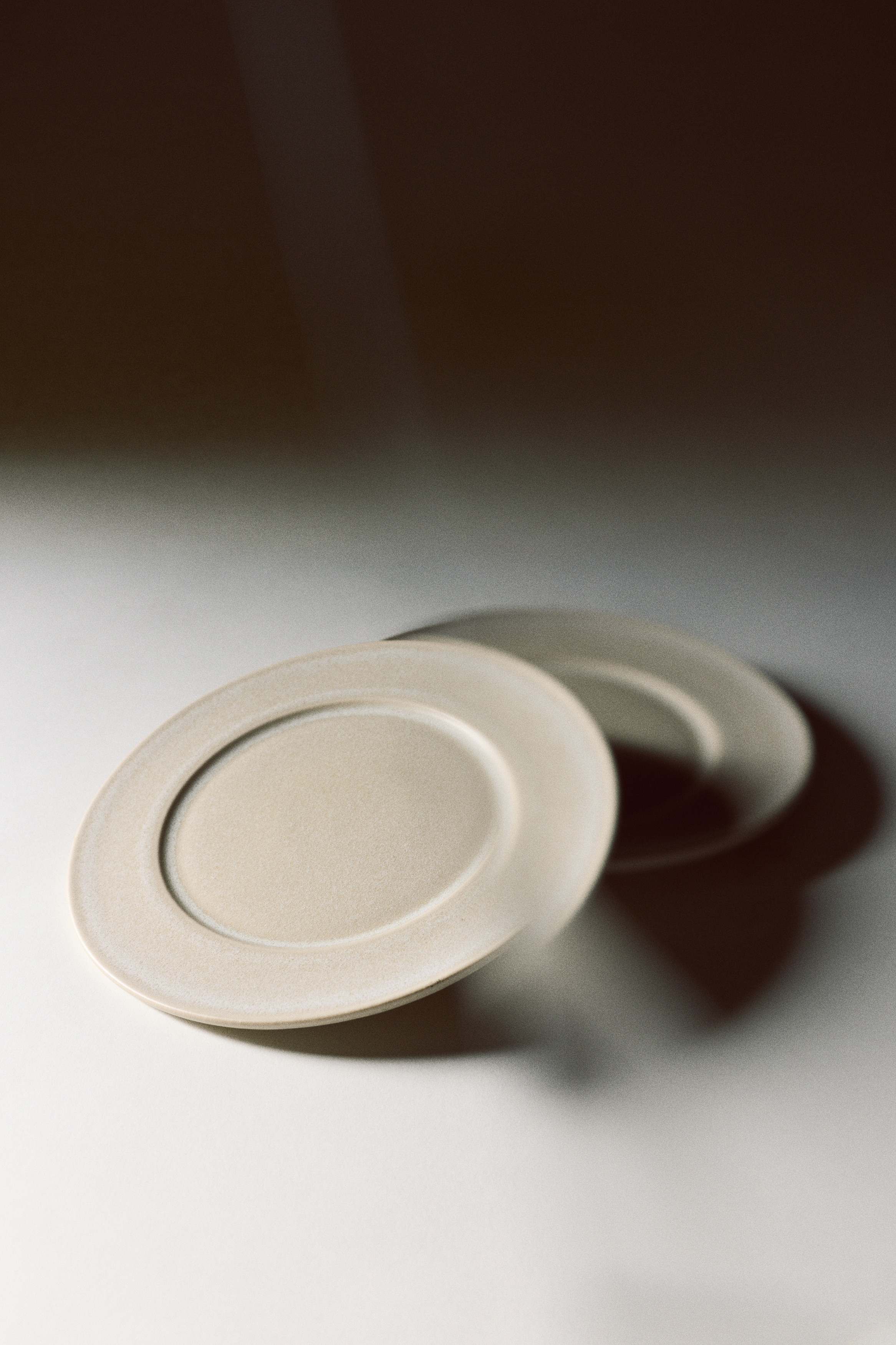 A round ceramic plate is lying flat in an overhead view, showcasing a textured, light-beige speckled glaze with a wide rim and a recessed circular centre.