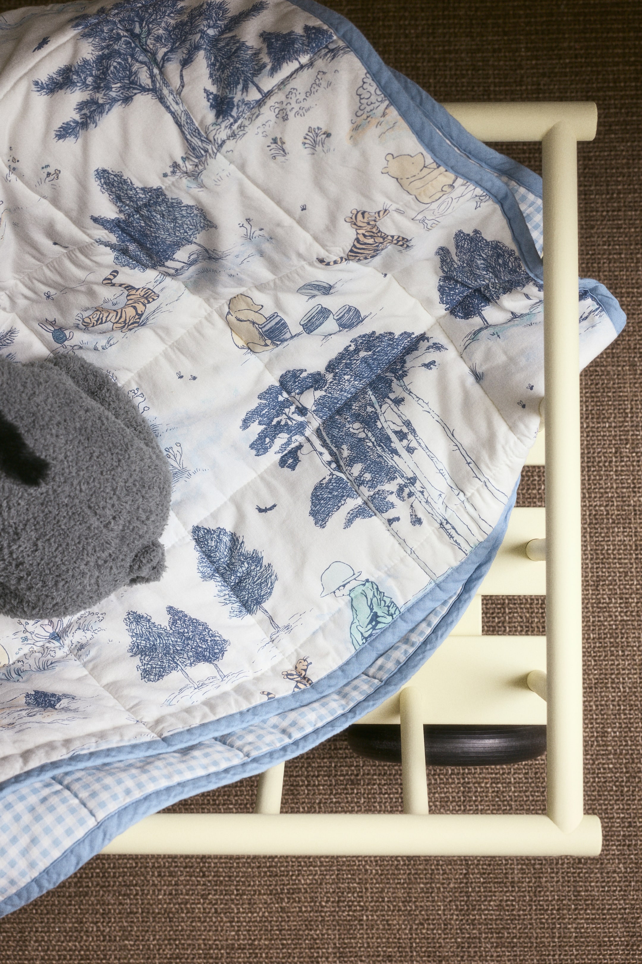 Näytä suurempi kuva: Tikkaillussa peitossa on sinivalkoinen toile-kuvio, jossa on puita ja Nalle Puh -hahmoja, vaaleansininen reunus ja vaaleansininen gingham-kääntöpuoli. Harmaa pehmolelu lepää peiton päällä, joka on aseteltu keltaisen pinnasängyn kaiteen yli.