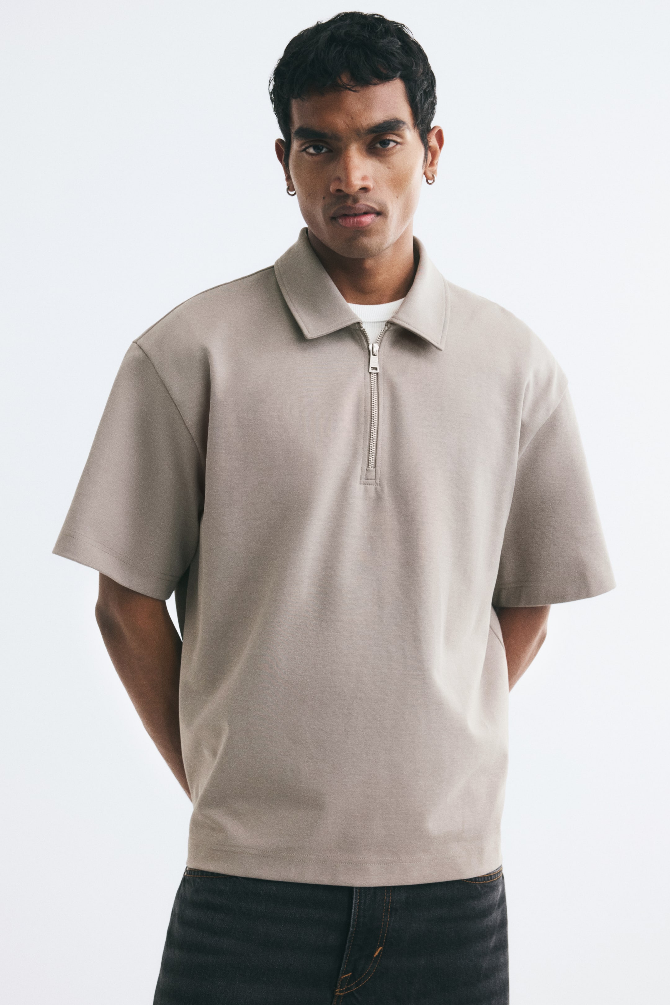 View larger image: A light taupe polo shirt in structured interlock cotton jersey is being worn by the model, featuring a relaxed fit, wide short sleeves, a classic collar, and a silver-toned half zip. The shirt is styled over a white T-shirt visible at the collar and is paired with dark jeans.