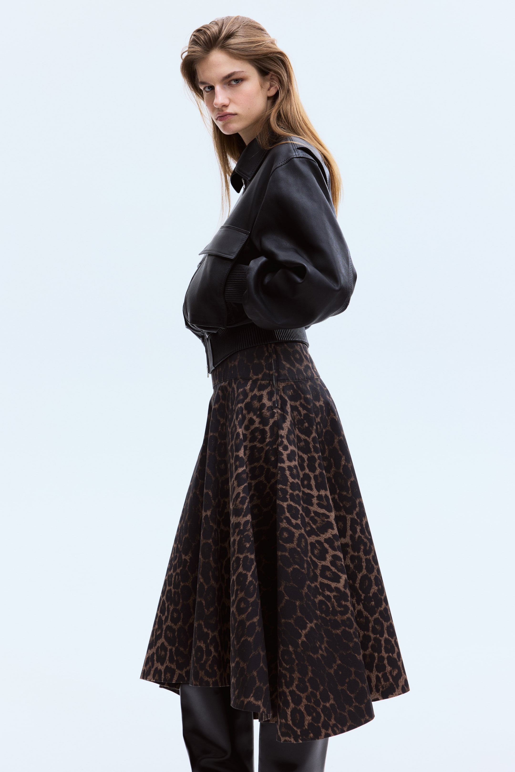 Visa större bild: En svart läderbomberjacka med spetsig krage, lockfickor, ribbade muddar och ribbad midjeband bärs över en utsvängd midikjol med ett brunt och svart leopardmönster. Jackan och kjolen är vinklade något åt vänster, vilket visar jackans voluminösa ärmar och kjolens fulla silhuett när den faller ovanför höga svarta stövlar.