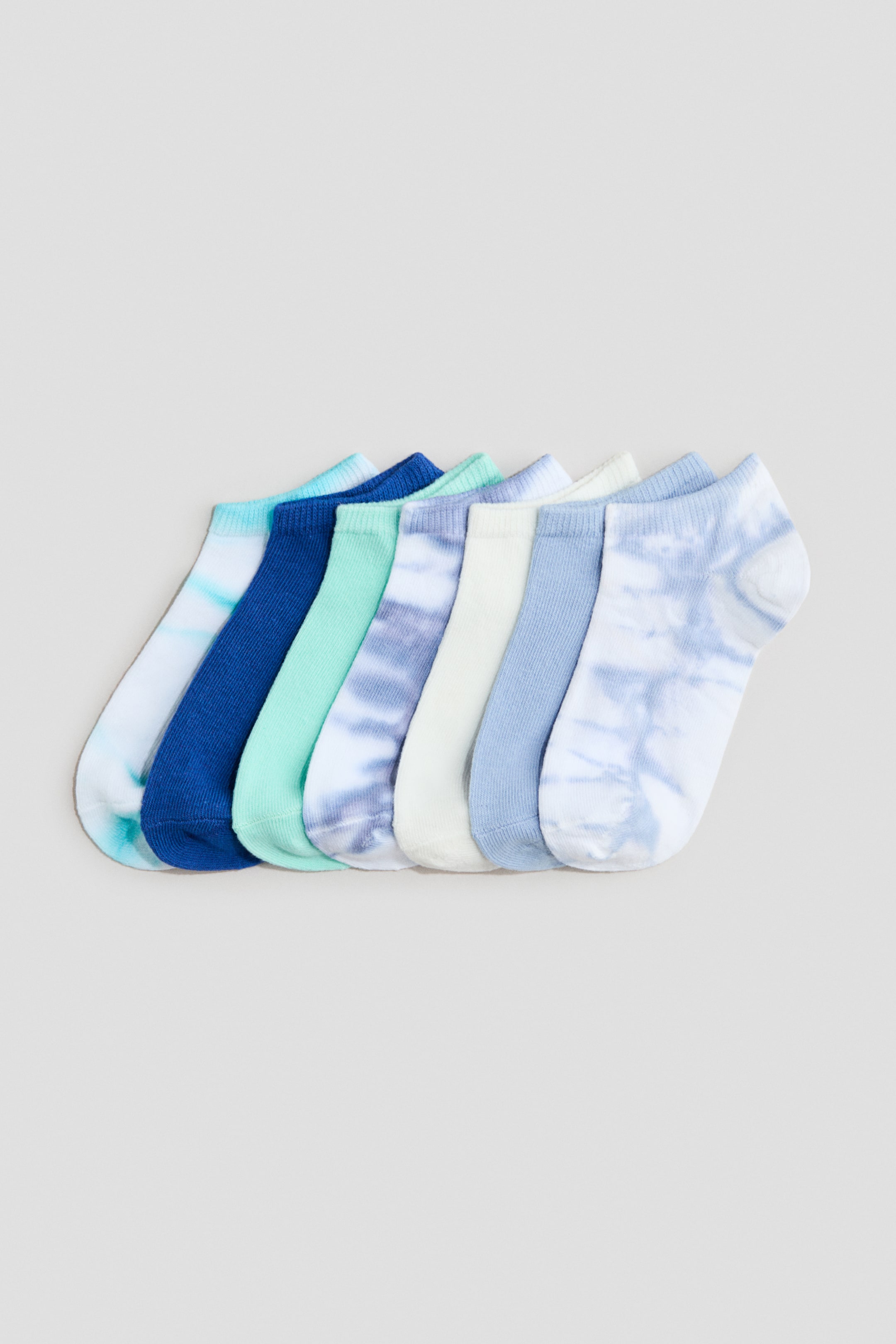 View larger image: Trainer socks are displayed in an overlapping line, highlighting seven pairs in a mix of solid and tie-dye patterns of blue, green, and white. Each sock boasts a fine-knit texture and narrow elastication at the top.