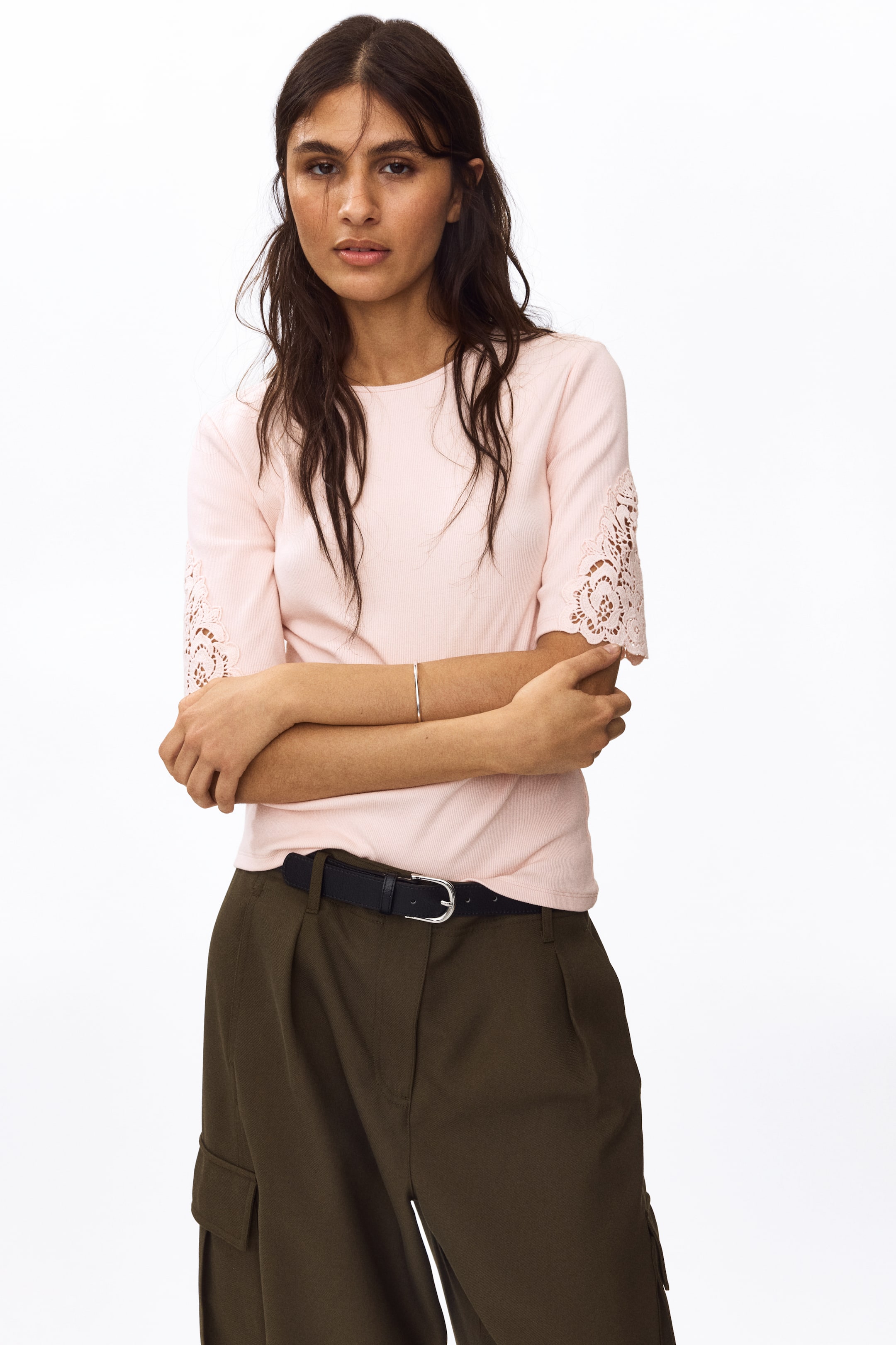 View larger image: The model is wearing a fitted top in a light pink ribbed cotton jersey, designed with a round neckline and short sleeves accented by lace insets. The top is neatly tucked into dark green trousers, held in place by a black belt, as the model stands confidently with arms crossed.