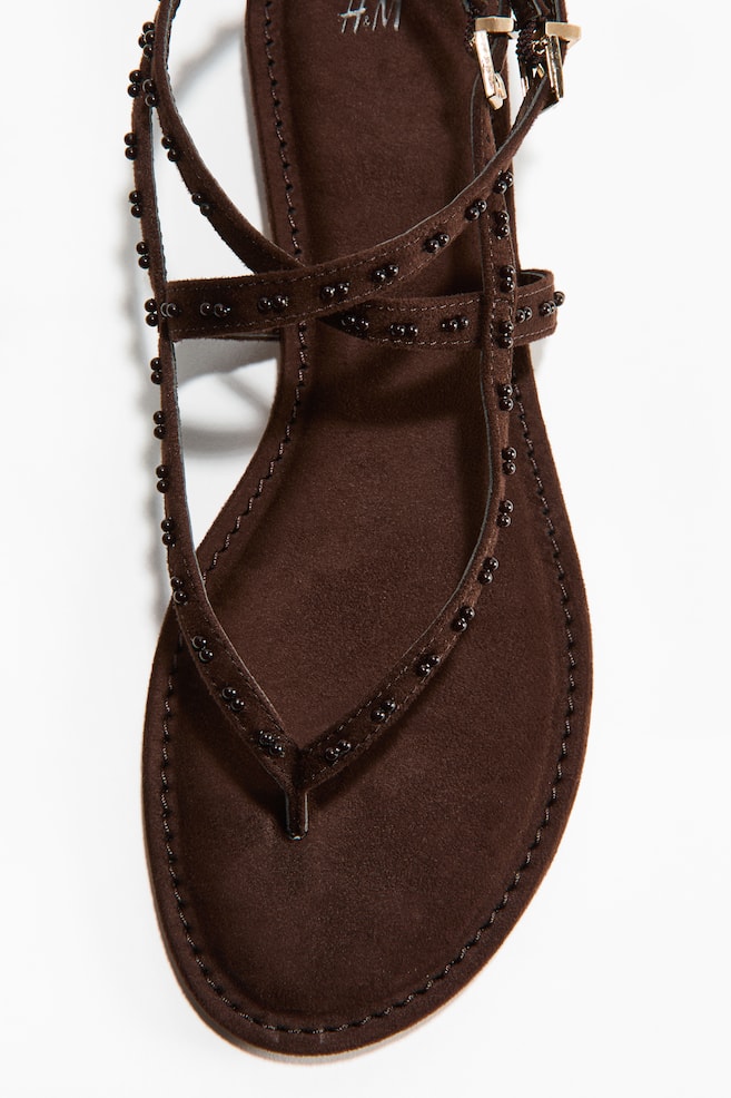 Une sandale marron présente un design à bride en T, avec des lanières ornées de petites perles rondes et sombres. Une bride de cheville avec une boucle dorée est visible au niveau du talon, et la semelle intérieure douce et marron montre des coutures visibles autour de son périmètre, avec le logo H&M imprimé sur le talon. - 1