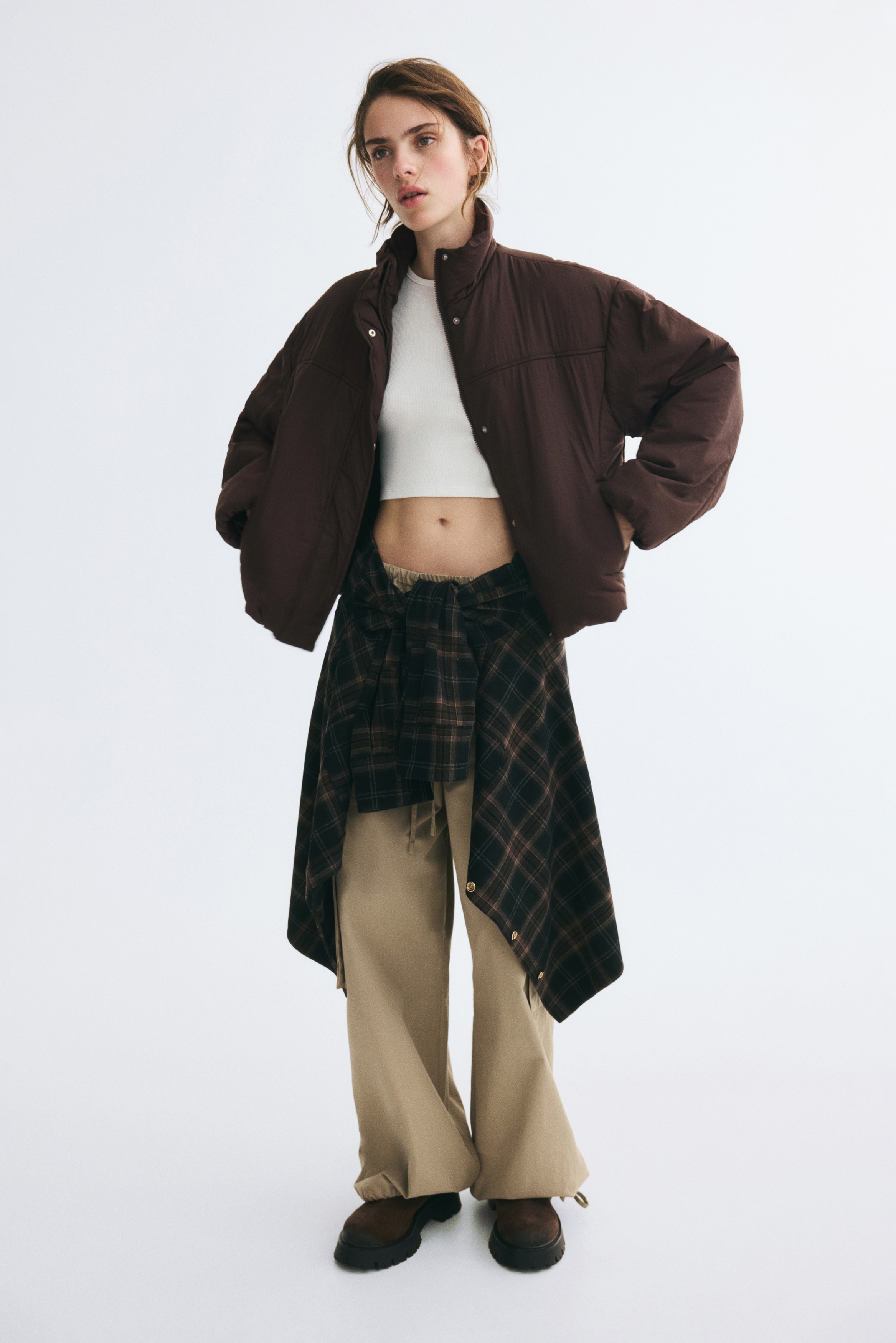 A dark brown puffer jacket is facing forward, featuring a high stand-up collar and a concealed front closure. The jacket showcases a padded construction with dropped shoulders, wide sleeves, side zippered pockets, and an elasticated hem.