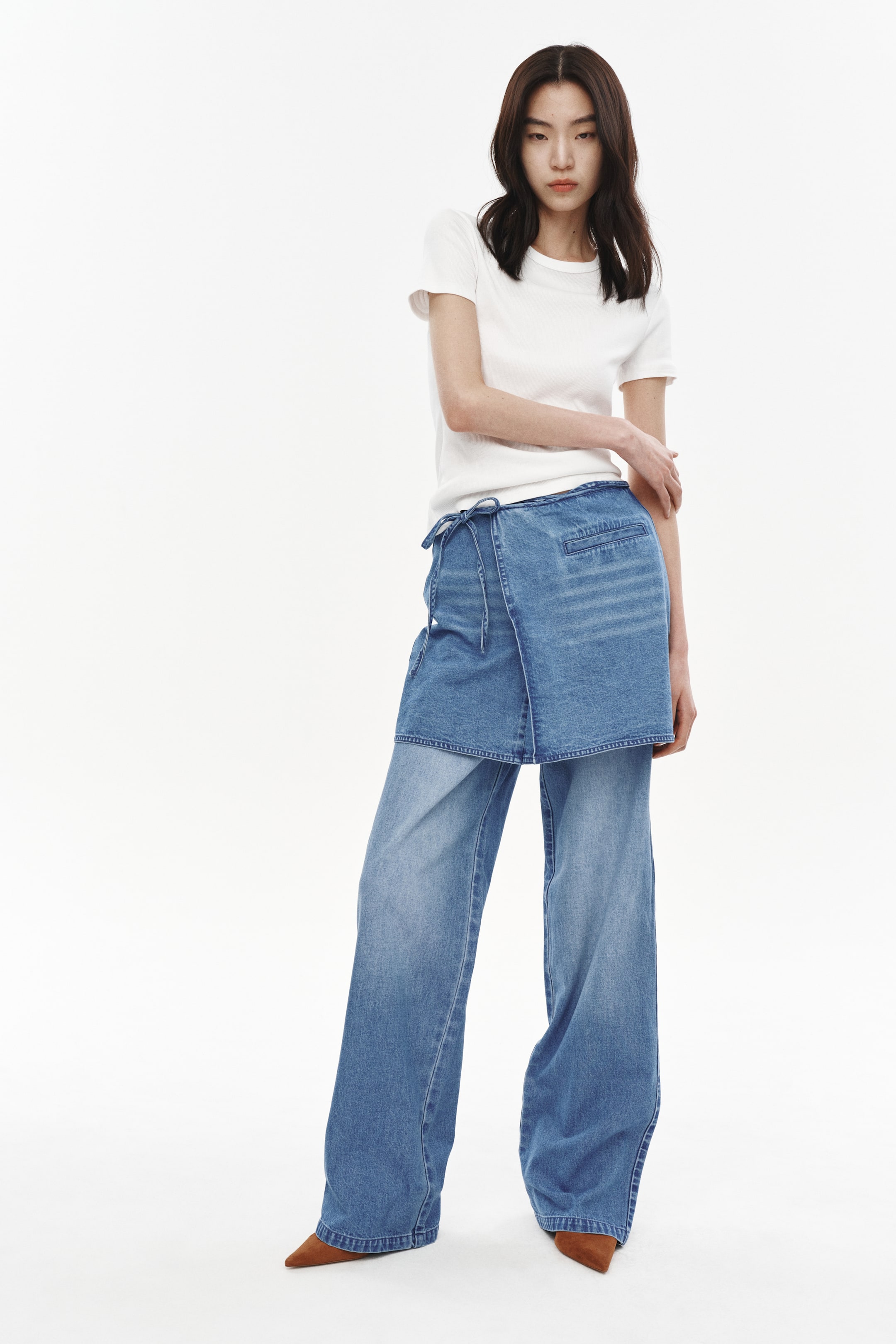 Xem ảnh lớn hơn: Quần jean denim ống rộng màu xanh nhạt được thiết kế với một lớp vải denim xếp lớp quanh eo, cố định bằng dây rút có thể nhìn thấy. Quần được trình bày hướng về phía trước, kết hợp với áo thun cổ tròn màu trắng và hơi xếp nếp trên đôi giày mũi nhọn màu nâu.