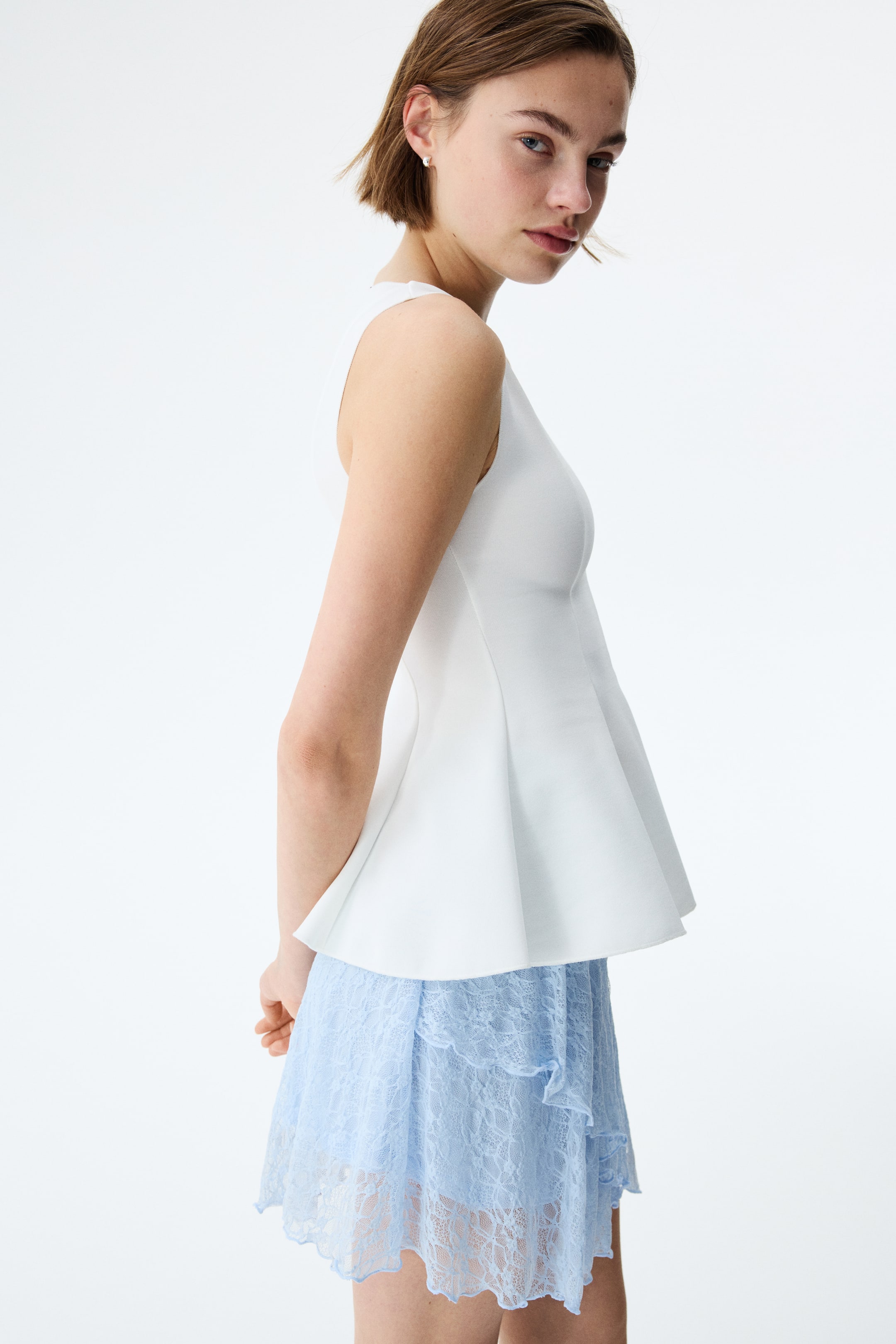 View larger image: The white sleeveless peplum top showcases a snug bodice that seamlessly transitions into a broad, structured flare at the waist, viewed from a subtle side angle. It is elegantly layered over a light blue lace mini skirt, which features ruffled tiers and a scalloped hem.