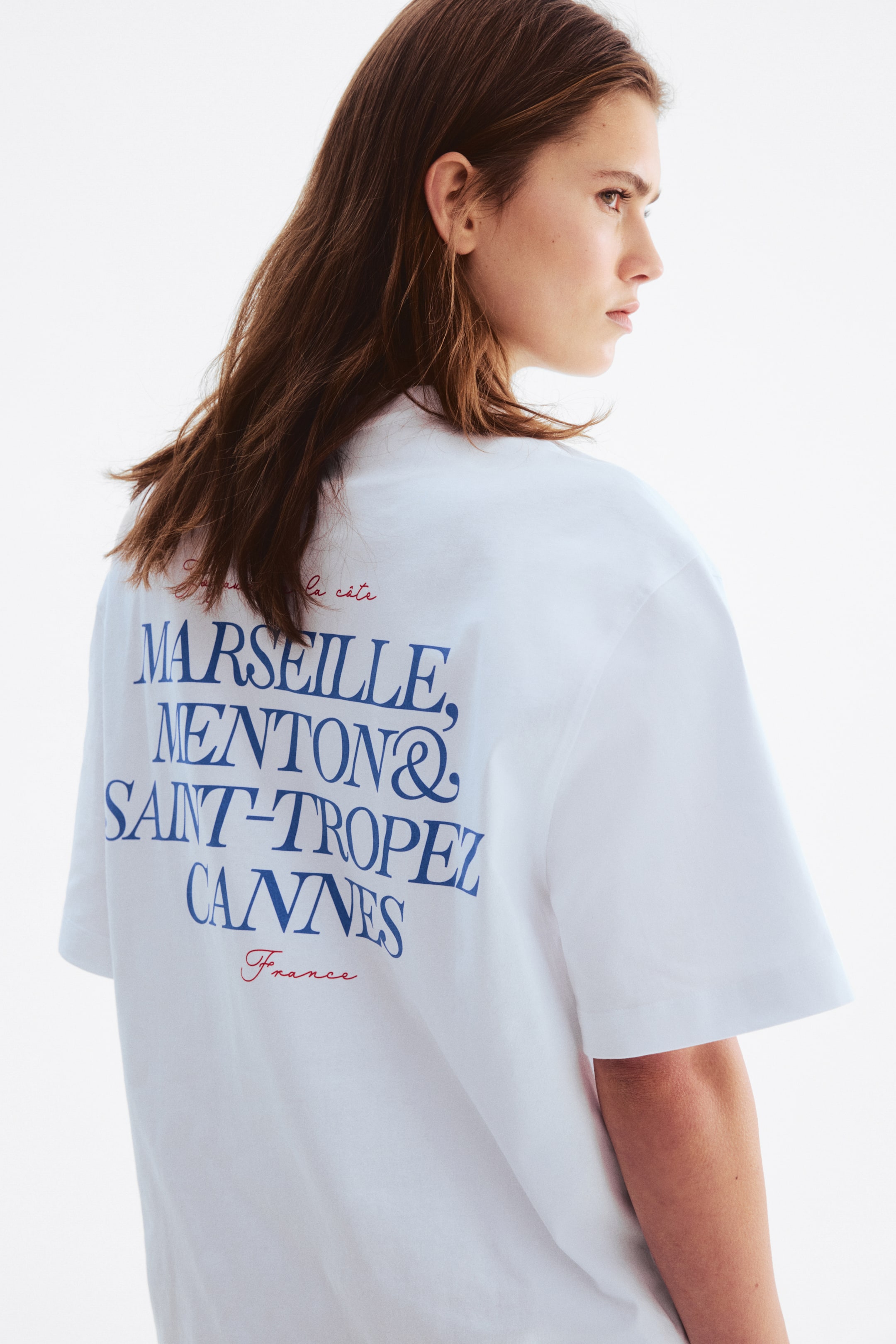Größeres Bild anzeigen: Ein weißes T-Shirt ist von hinten zu sehen und hat kurze Ärmel. Es zeigt großen blauen Drucktext mit der Aufschrift "MARSEILLE, MENTON & SAINT-TROPEZ CANNES," darüber steht in kleinerem roten Text "la côte" und darunter "France."