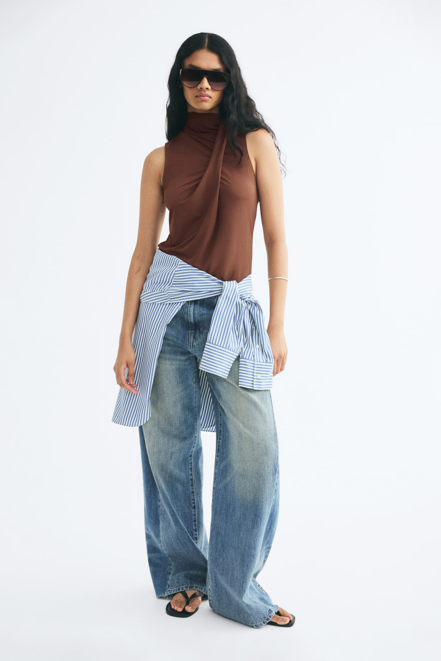 A brown sleeveless top in stretch jersey is worn by the model, featuring a high stand collar that flows into a draped front panel. The top is styled with wide-leg blue jeans and a blue-and-white striped shirt tied around the waist. - 1