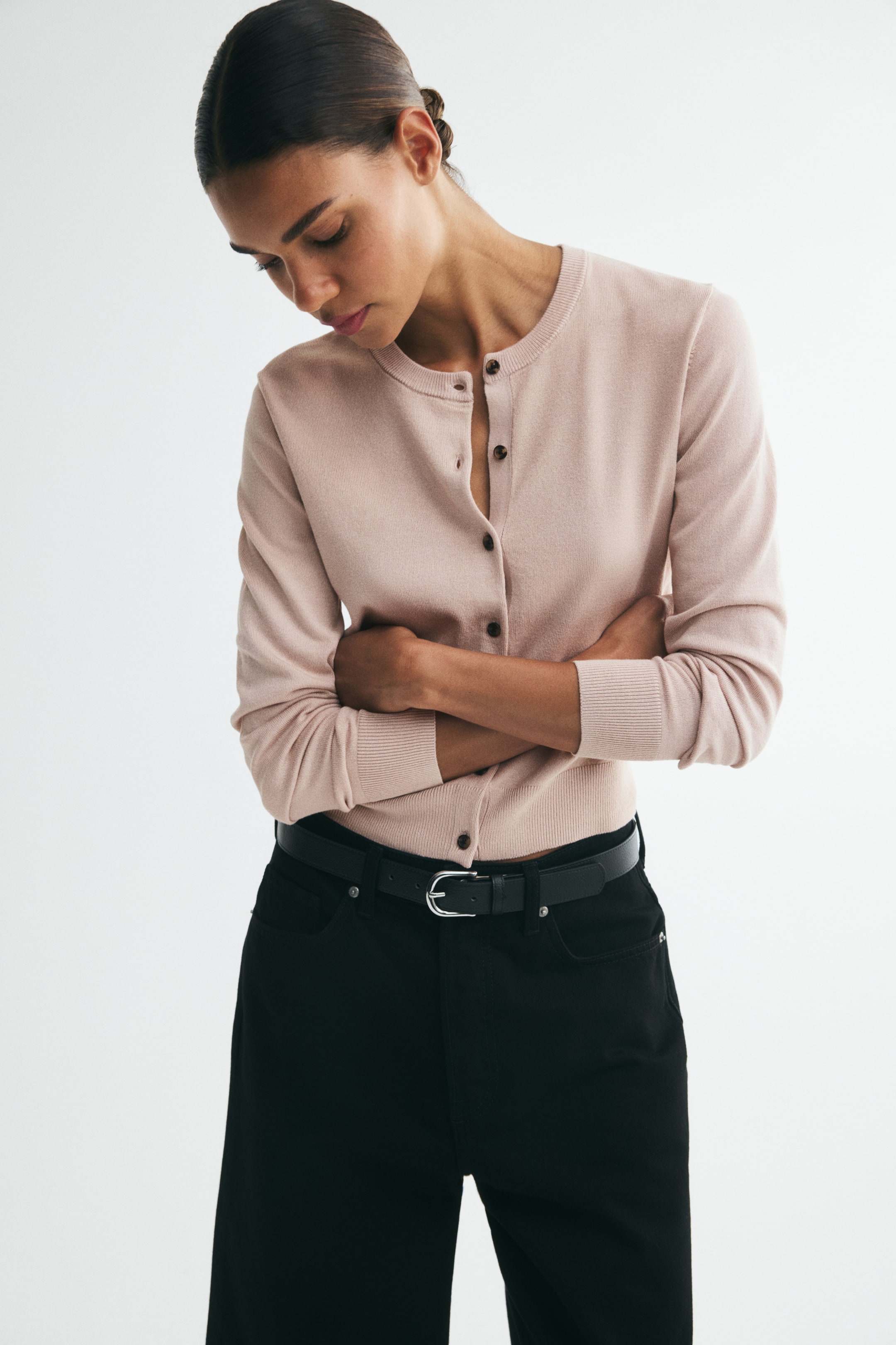 Größeres Bild ansehen: Ein rosa Feinstrick-Cardigan mit Rundhalsausschnitt, Knopfleiste vorne sowie gerippten Bündchen und Saum wird von dem Model getragen. Einige Knöpfe sind vorne offen, und das Model hat die Arme verschränkt, während sie ihn über einer schwarzen Hose mit Gürtel trägt.