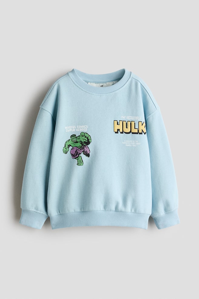 Un pulover fleece bleu deschis este prezentat din faÈÄ, evidenÈiind un decolteu rotund cu margini striate, umeri cÄzuÈi Èi manÈete Èi tiv striate. Ãn zona pieptului este afiÈat un grafic imprimat cu personajul Hulk pe o parte Èi textul âTHE INCREDIBLE HULKâ cu litere groase galbene pe cealaltÄ. - 1