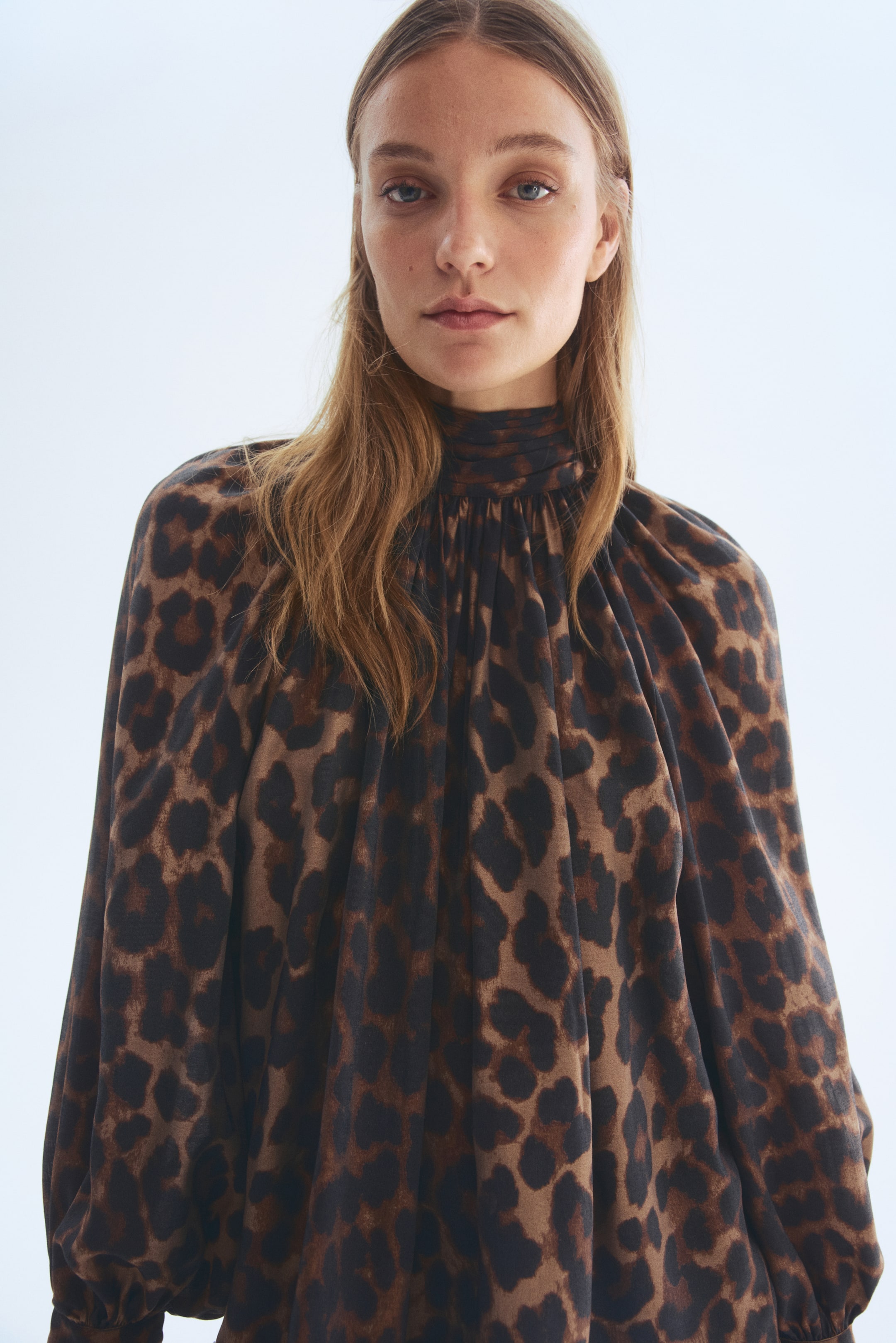 Nagyobb kép megtekintése: Egy leopárdmintás blúz, amely magas, ráncolt nyakkivágással és bő, hosszú ujjakkal rendelkezik, amelyek a mandzsettáknál gyűlnek össze, laza, áramló illeszkedést biztosítva elölről nézve.