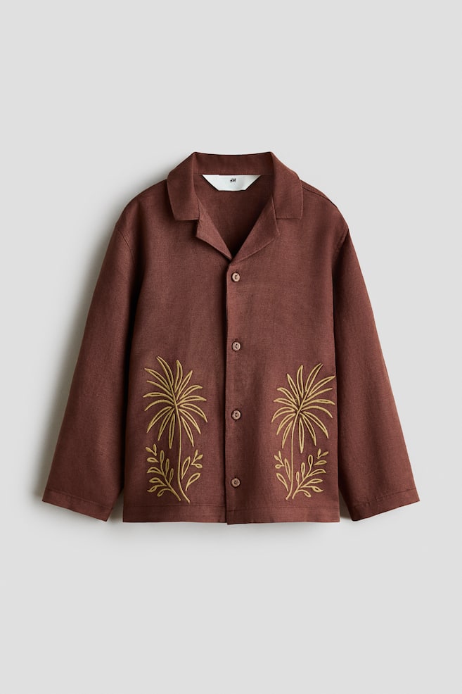 A brown, linen-textured long-sleeve shirt is shown from the front, featuring a camp collar and a button-down closure. Two yellow-gold embroidered palm tree motifs detail the lower left and right front panels. - 1