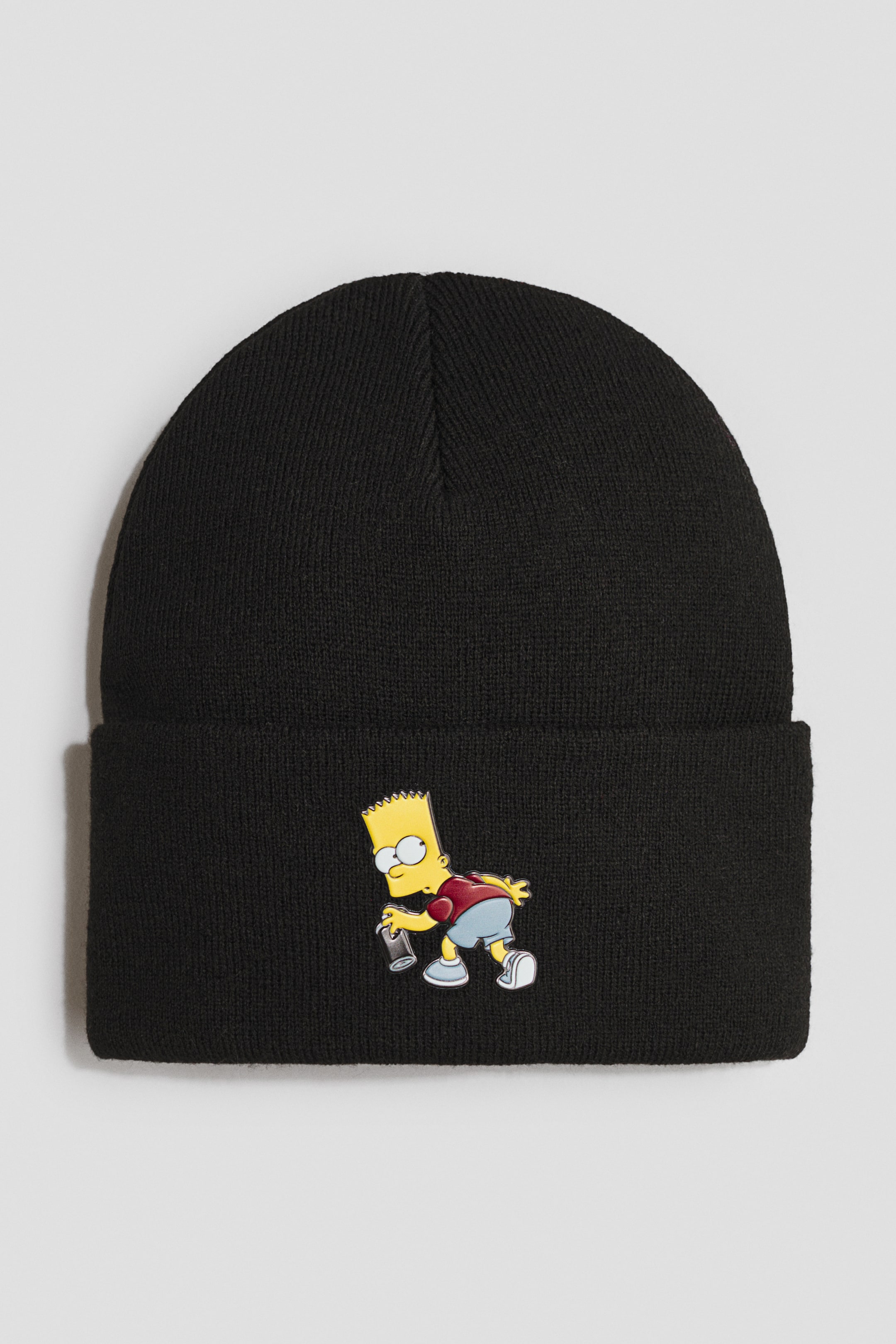 Größeres Bild ansehen: Eine schwarze Strickmütze zeigt auf dem umgeschlagenen Bund auffällig eine farbenfrohe Grafik von Bart Simpson, der eine Sprühdose hält, und präsentiert ihre runde Krone sowie die sichtbare Strickstruktur.