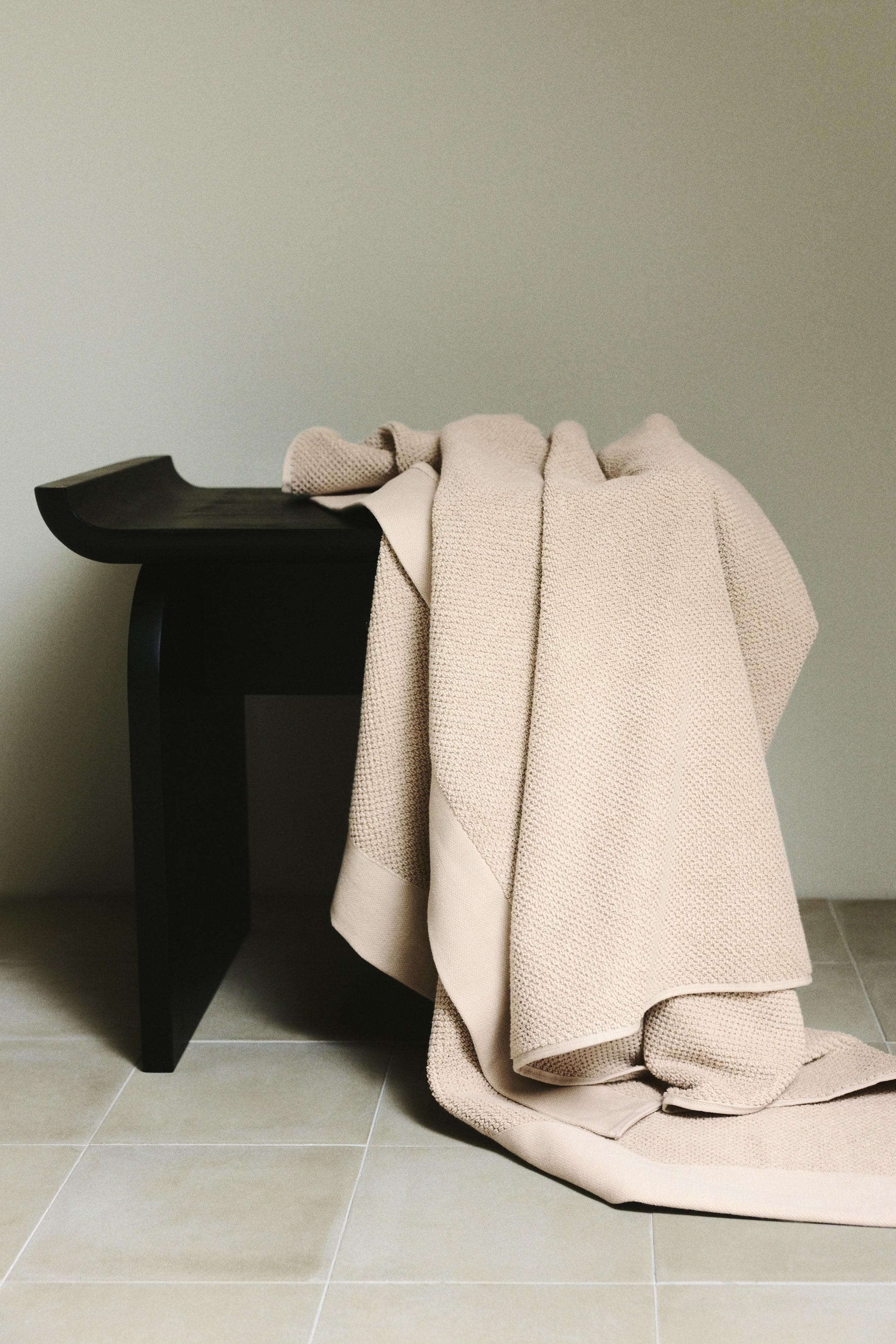 View larger image: A light beige bath towel is draped, featuring a textured waffle-knit surface and smooth, flat borders along its edges.