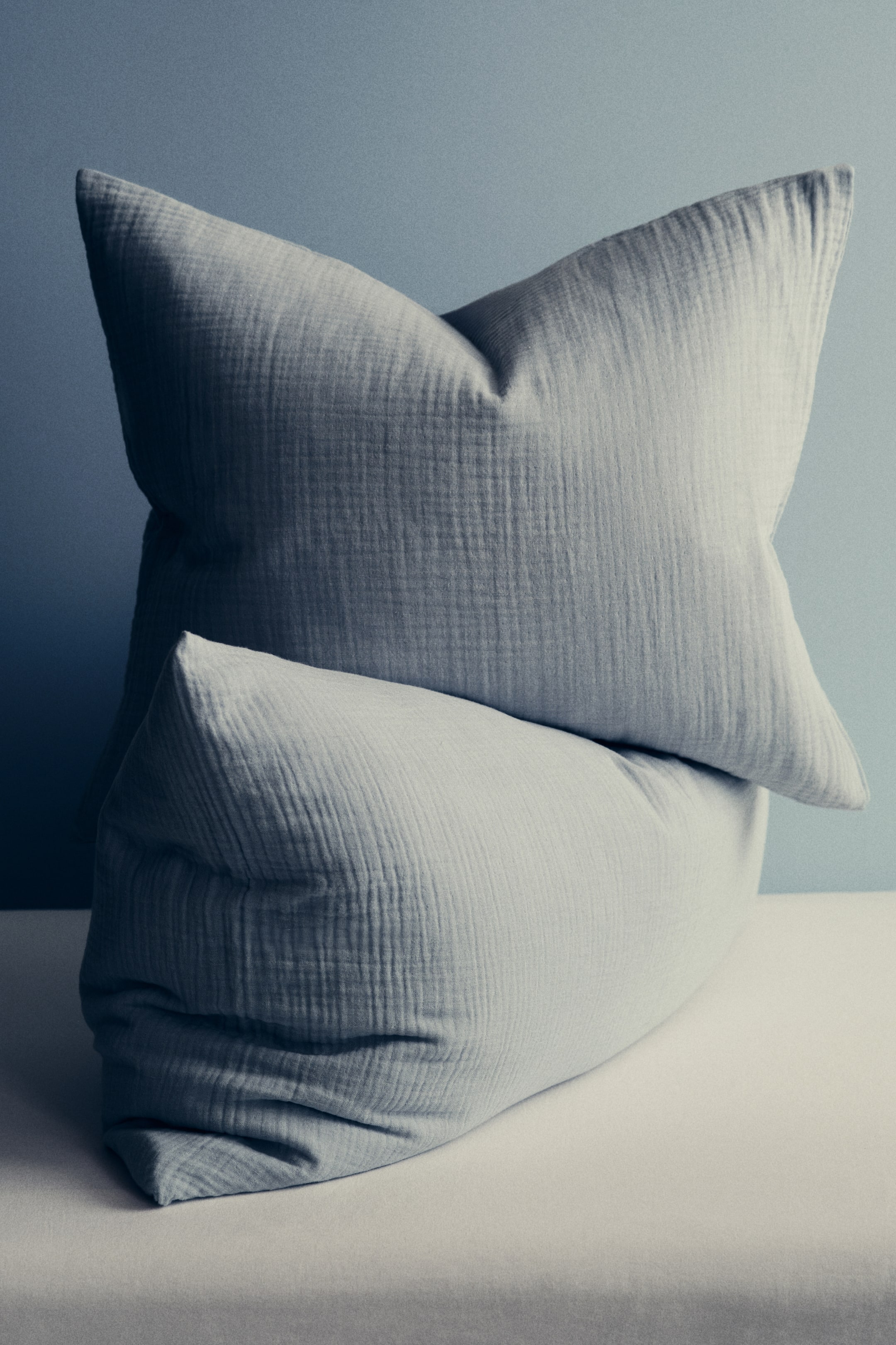 Agrandir l'image: Deux coussins gris clair mettent en valeur un tissu en mousseline texturée. Le coussin supérieur, bien rebondi, est dressé verticalement, incliné légèrement vers la gauche, tandis que le coussin inférieur repose à l'horizontale devant, incliné légèrement vers la droite.