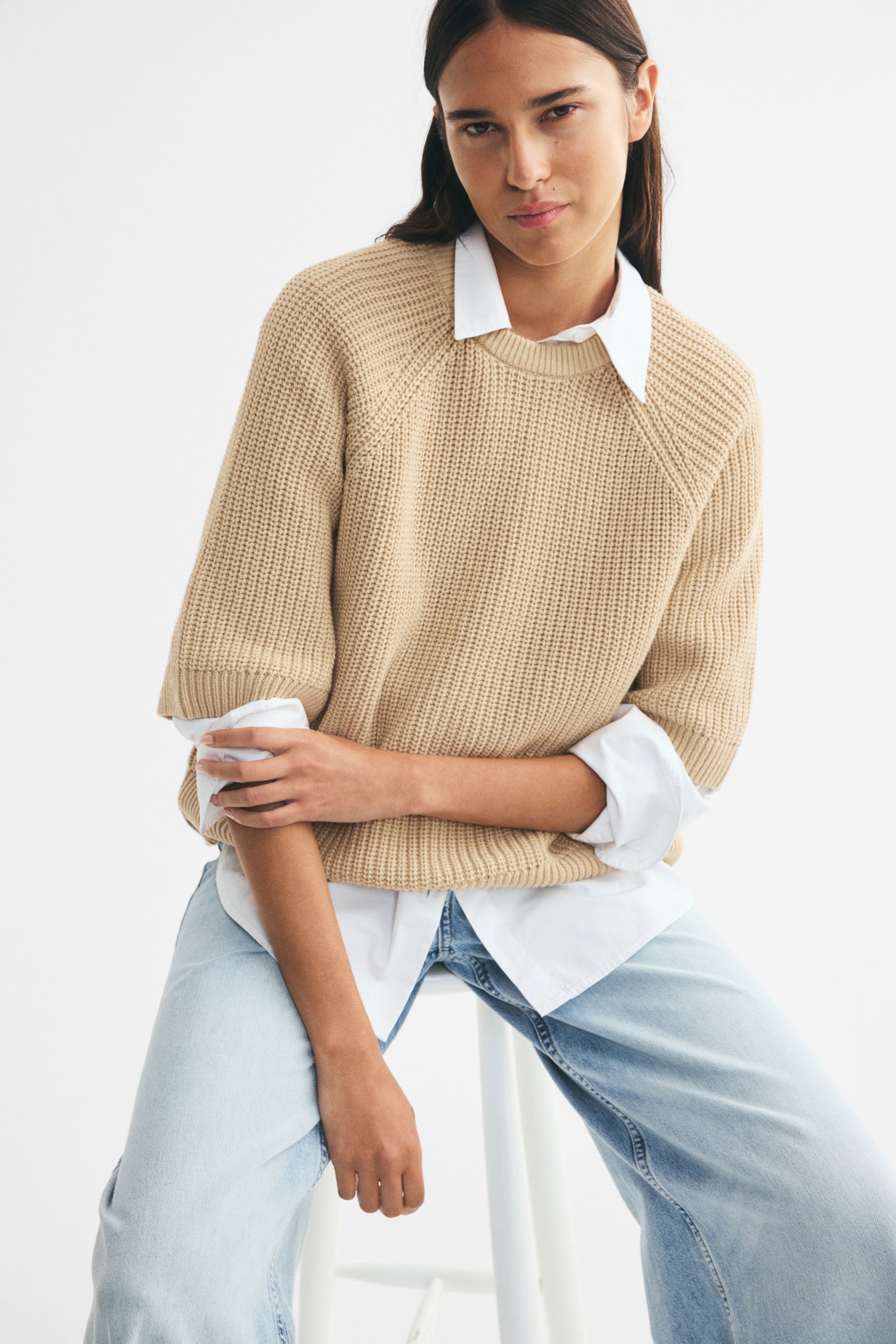 View larger image: The model is wearing an oversized beige rib-knit T-shirt with a round, rib-trimmed neckline and elbow-length raglan sleeves. It is layered over a white collared shirt and paired with light-wash jeans while the model is seated.