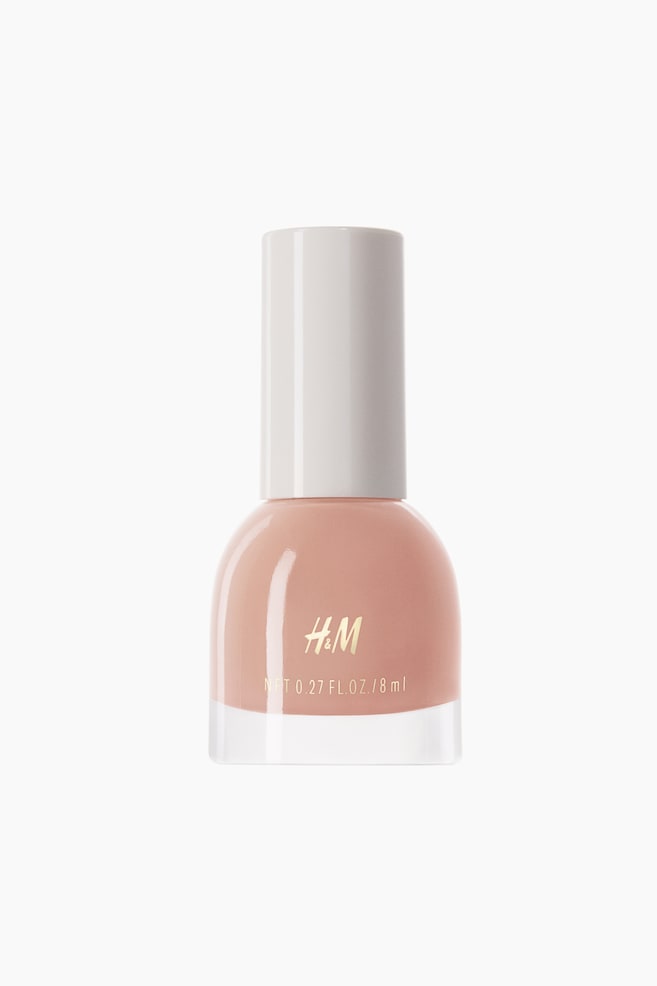 Um frasco de verniz exibe um verniz de unhas em tom pêssego claro. Apresenta um recipiente de vidro transparente com um ombro arredondado, uma tampa cilíndrica branca e o logótipo dourado "H&M", juntamente com detalhes de volume impressos na sua superfície frontal. - 1