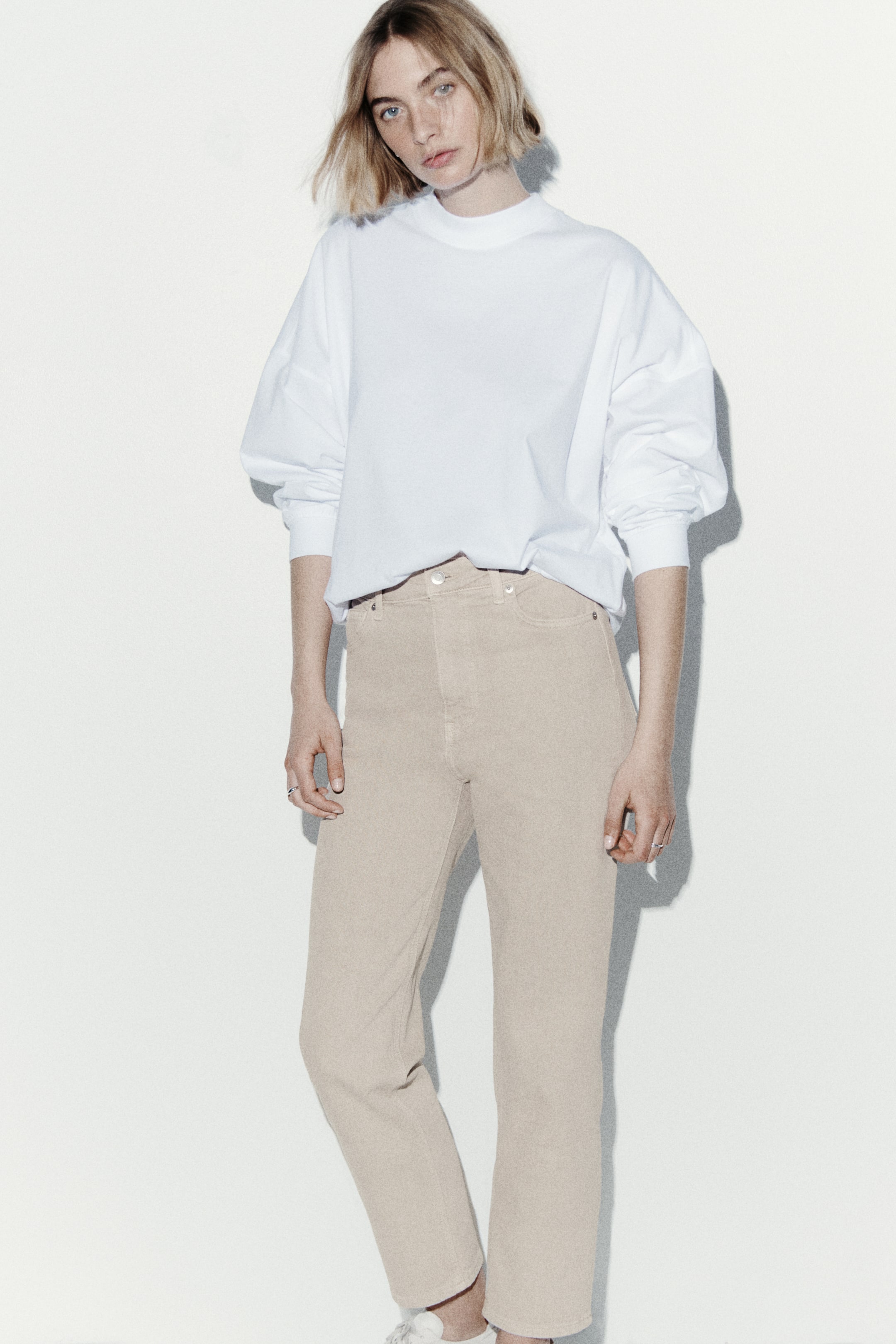 拡大画像を表示: ベージュのハイウエストジーンズは、ストレートレッグカットとアンクルレングスの裾が特徴で、フロントボタンとジッパークロージャーが目立ち、フロントポケットが付いています。白いスウェットシャツがウエストバンドに少しタックインされており、ハイライズフィットを強調しています。