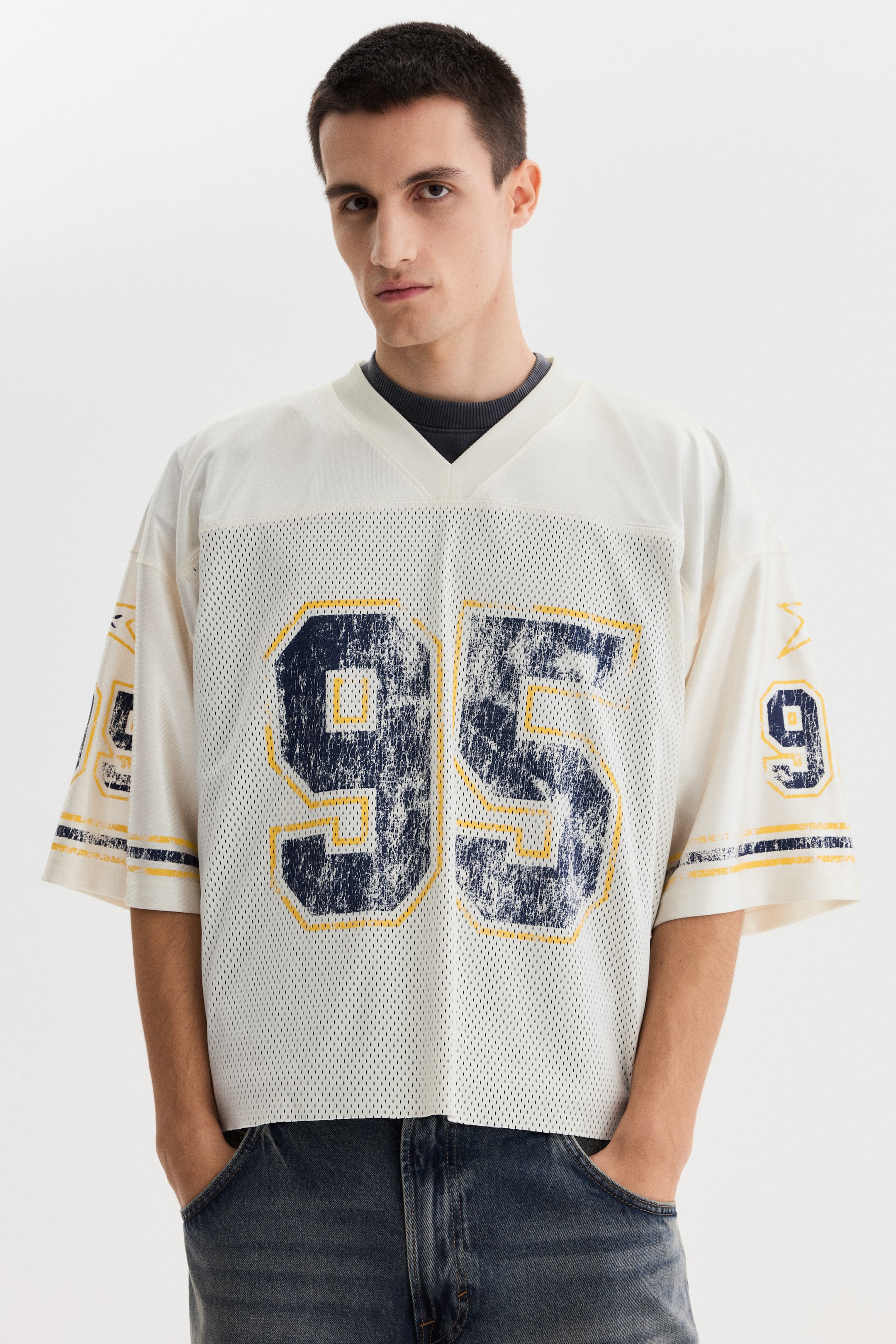 View larger image: An off-white mesh jersey features a ribbed V-neck and large, distressed dark blue numbers "95" outlined in yellow on the front. Distressed dark blue and yellow striped bands appear on the short sleeves, with a smaller distressed "9" and a yellow symbol visible on the right sleeve. The oversized jersey is worn over a dark grey crewneck sweater.