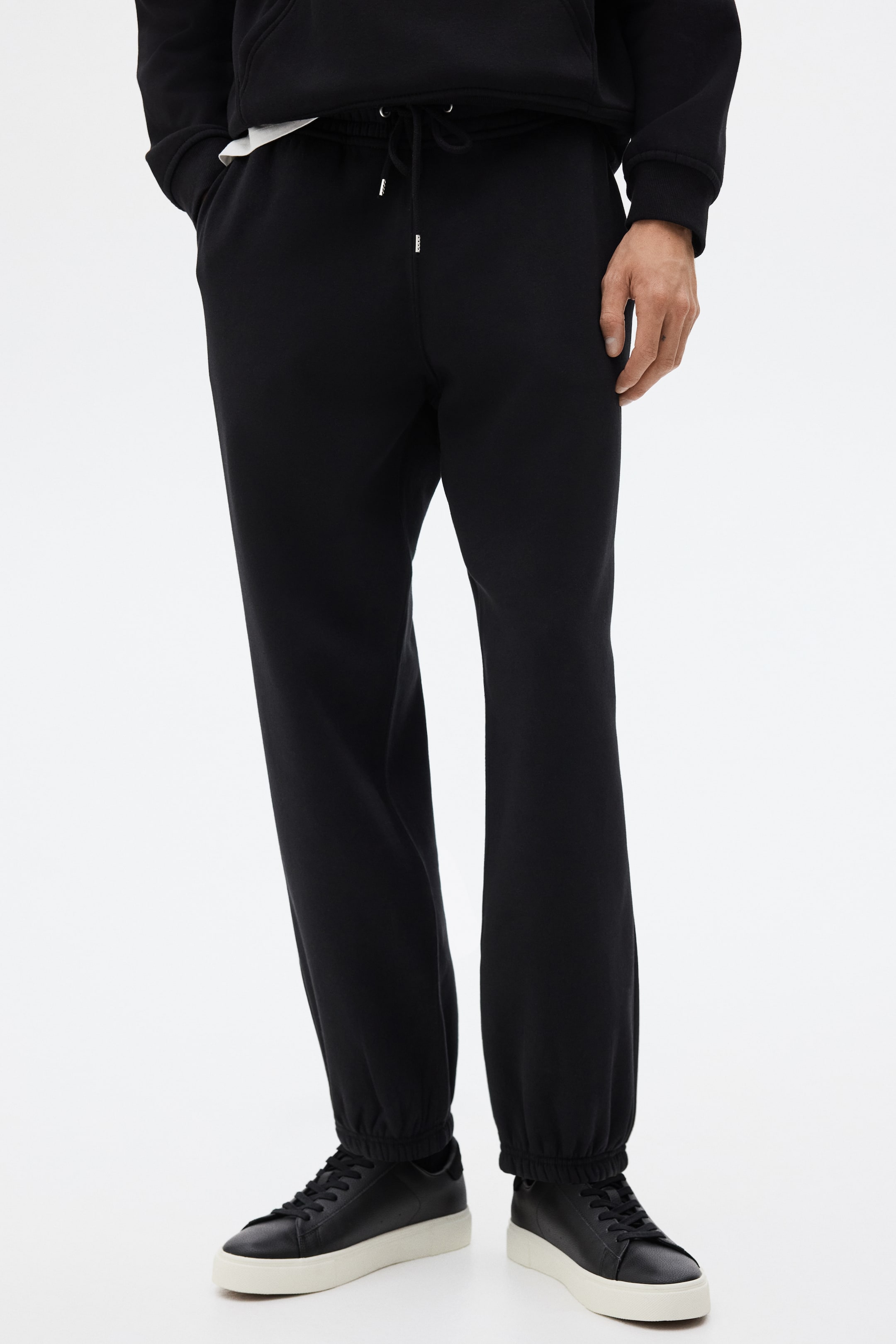 Visualizza immagine più grande: I pantaloni della tuta neri vengono mostrati da una vista frontale leggermente angolata, caratterizzati da una vita con coulisse elastica e aglet tono argento e tasche laterali. Le gambe sono tagliate con una vestibilità rilassata, raccogliendosi alle caviglie con polsini elasticizzati.