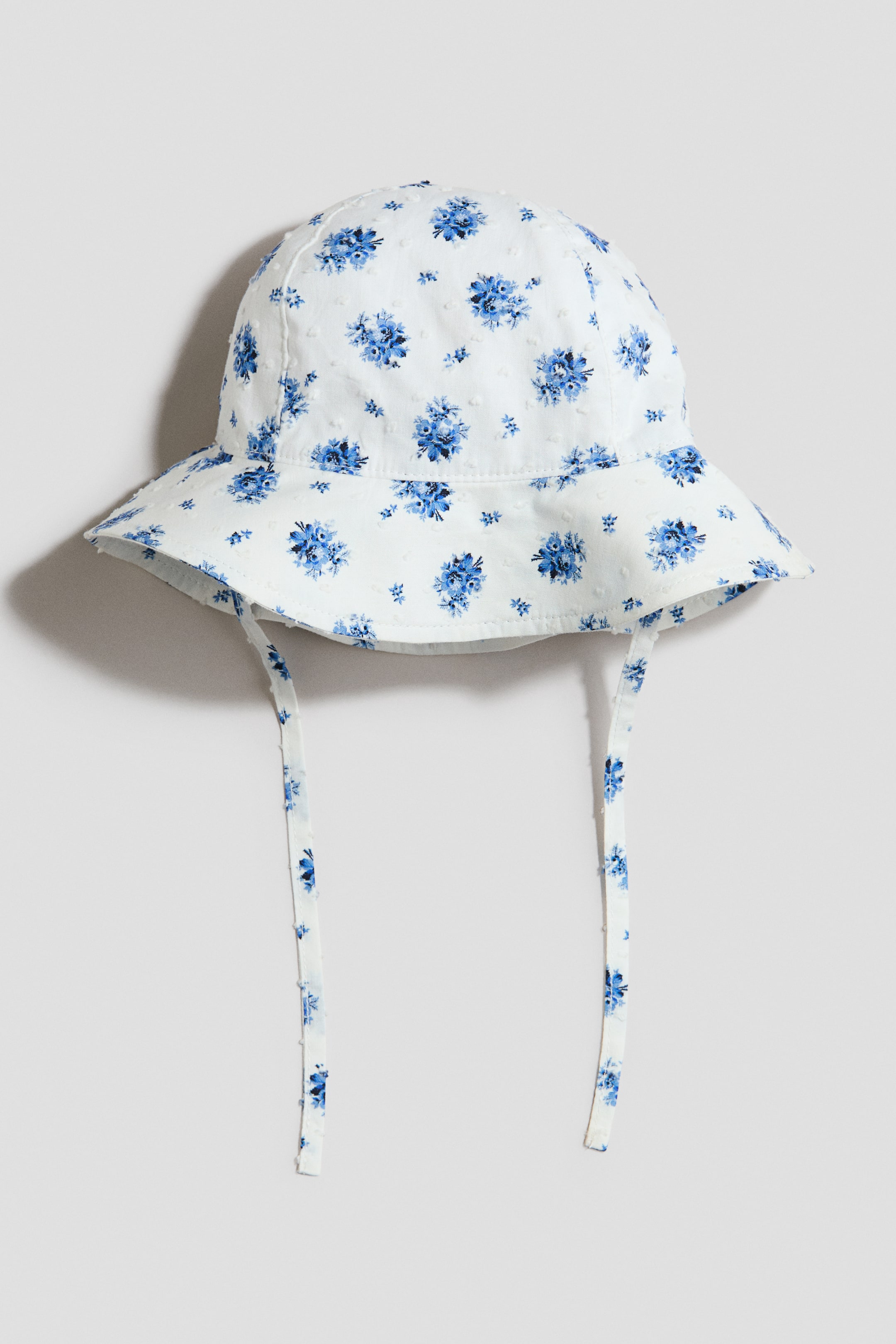 View larger image: "A white sun hat, featuring a blue floral print with raised dot detailing on the fabric, is displaying a soft, downward-sloping brim and two matching floral tie straps hanging from the front."
