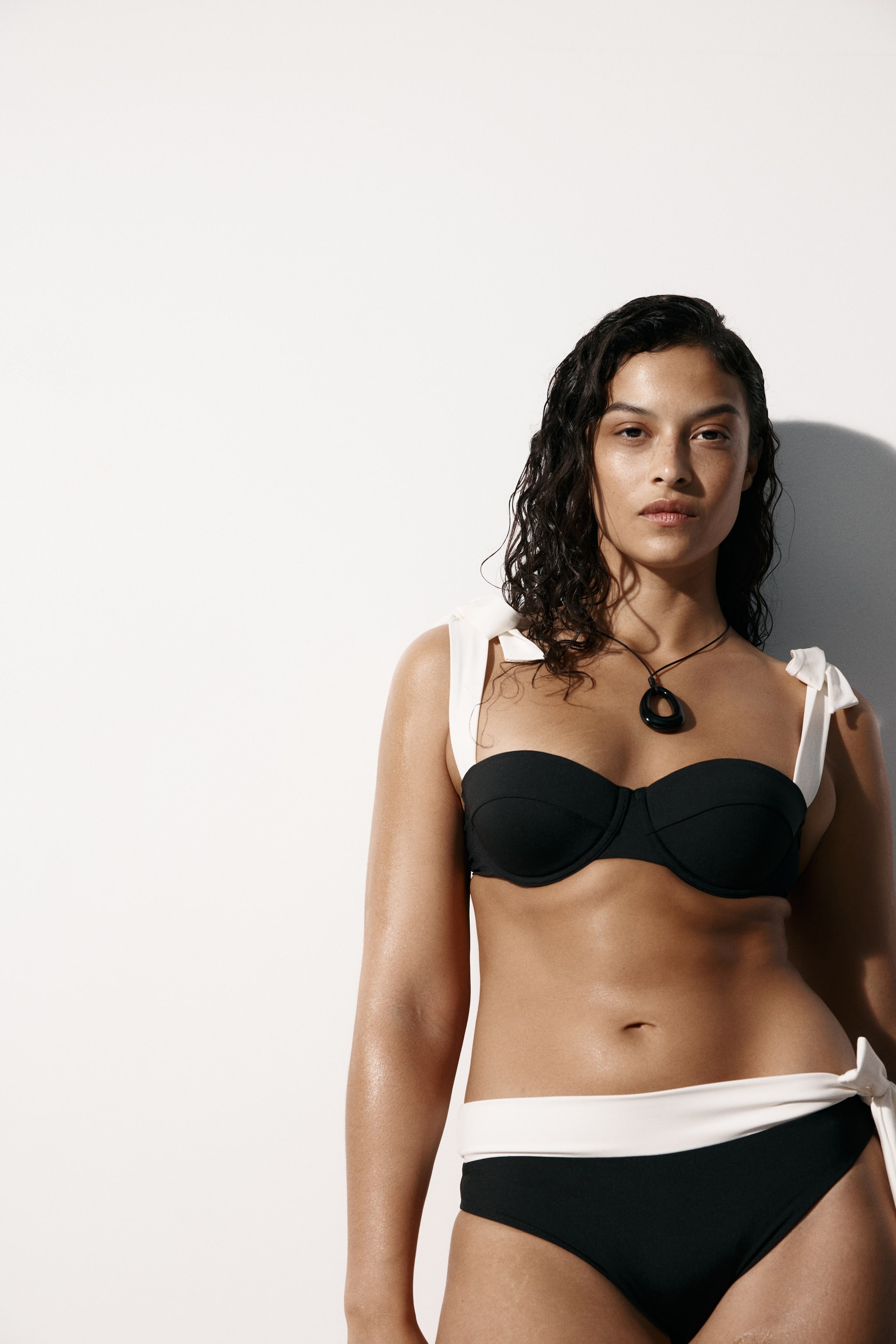 Ver imagen más grande: La modelo lleva un bikini de dos piezas. Presenta un top bandeau negro con tiras blancas en los hombros, combinado con braguitas negras que tienen una amplia banda blanca que se envuelve y ata al costado.