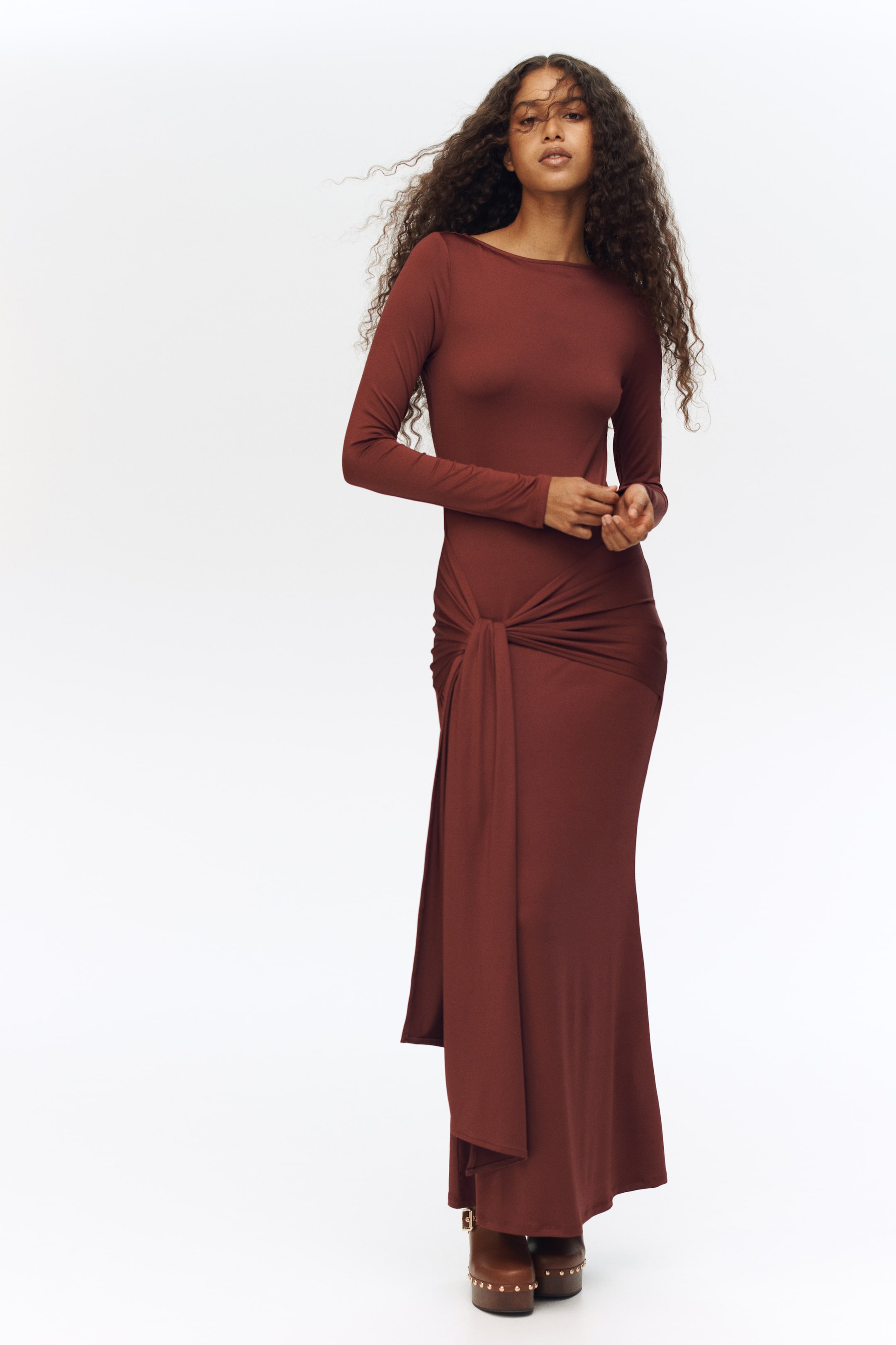 View larger image: A front-facing, rust-brown maxi dress features a boat neck and long sleeves, with a form-fitting silhouette. The dress is cinched at the left hip with a knot detail, creating a draped front panel and a side slit, flowing gracefully to a maxi length.