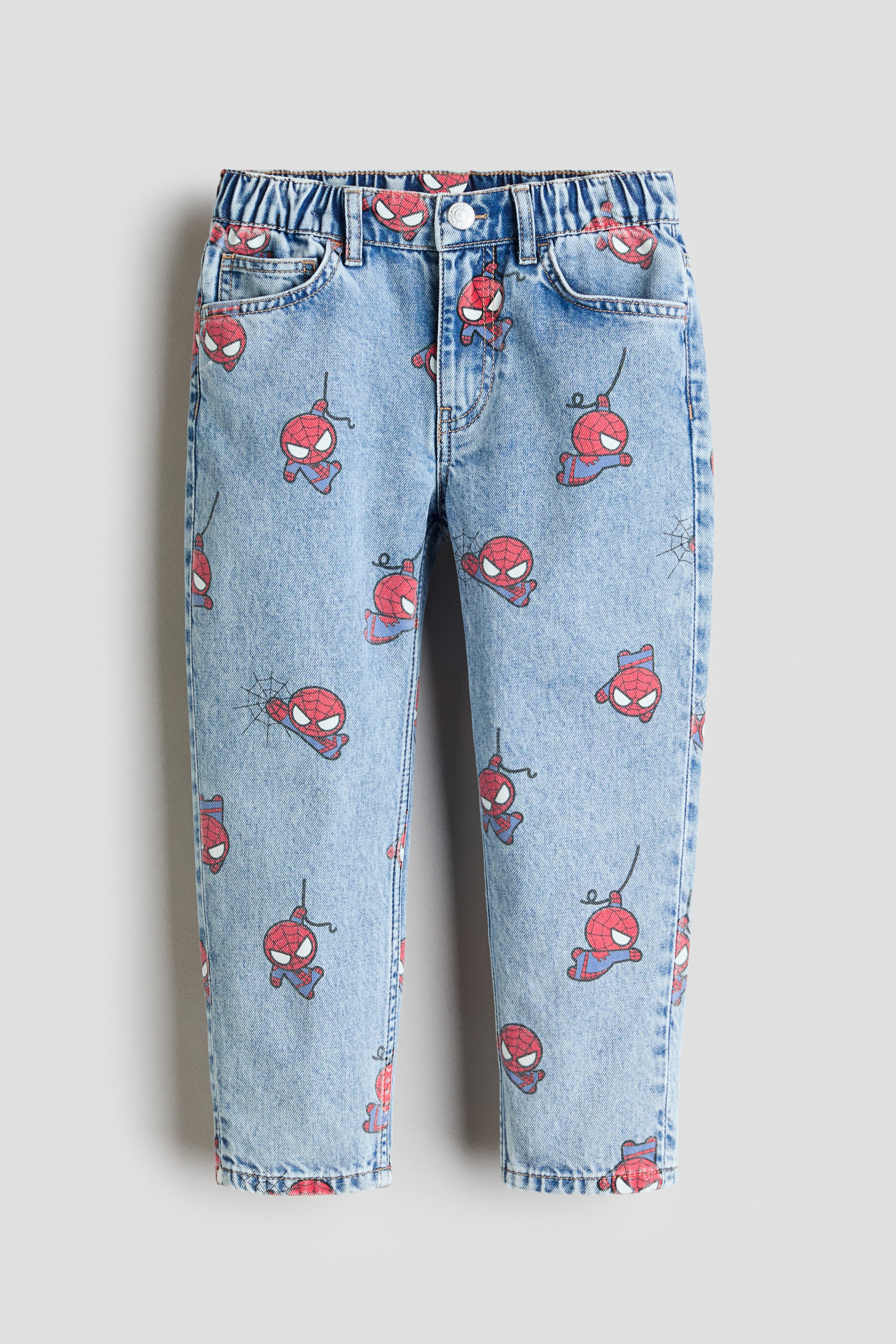 Ver imagen más grande: Estos vaqueros de 5 bolsillos se presentan en una vista frontal y están confeccionados en denim de algodón suave de color azul claro. Presentan un estampado integral de figuras de Spider-Man en rojo y azul, una cintura elástica con cierre de cremallera y botón, y un ajuste regular en la cadera y el muslo con piernas cónicas.