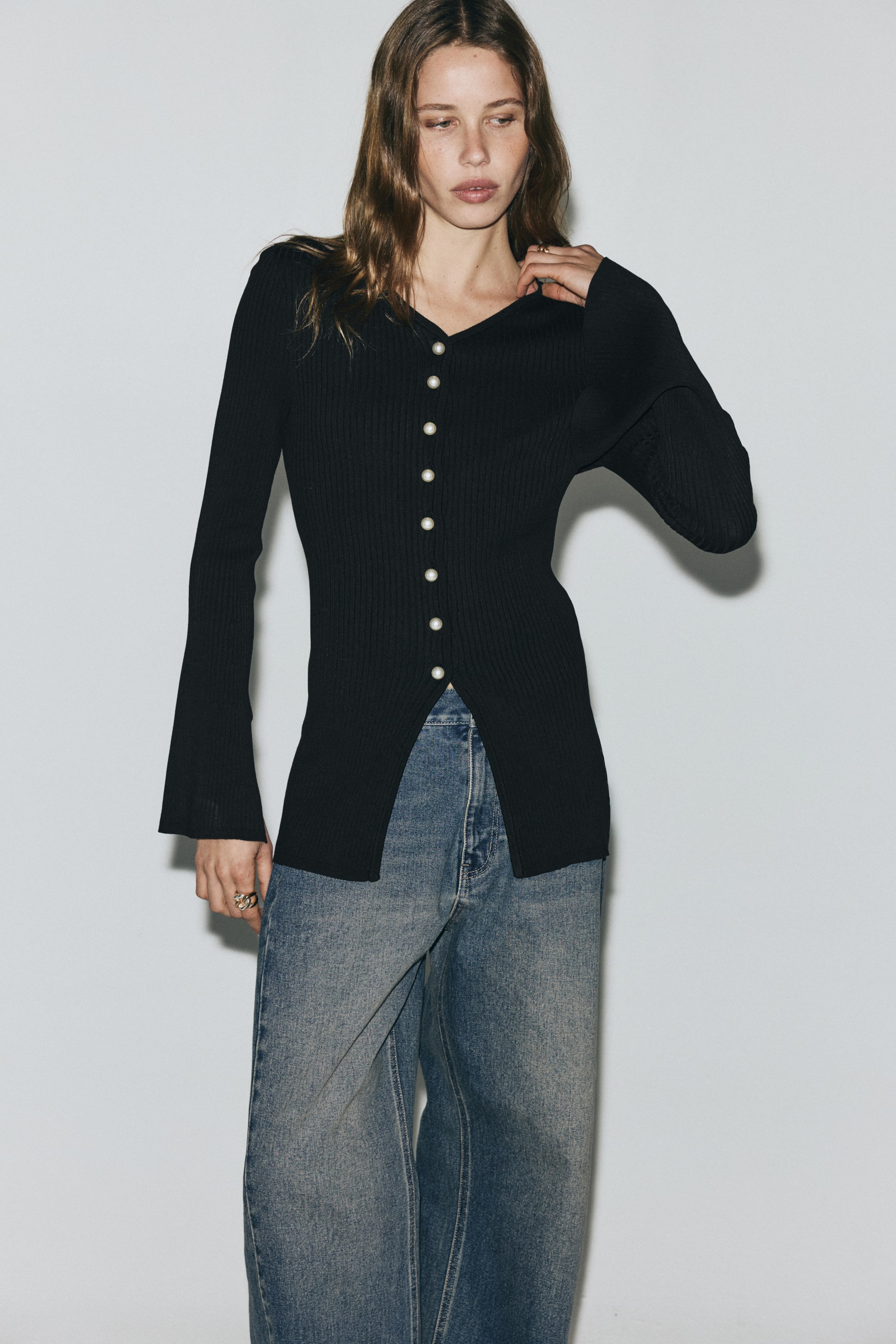 Visualizza immagine più grande: Un cardigan nero a coste è rivolto in avanti, caratterizzato da uno scollo a V e una chiusura a bottoni completa con bottoni simili a perle. Le maniche lunghe terminano con polsini svasati e il fondo del cardigan presenta uno spacco frontale poiché i bottoni inferiori sono sbottonati.