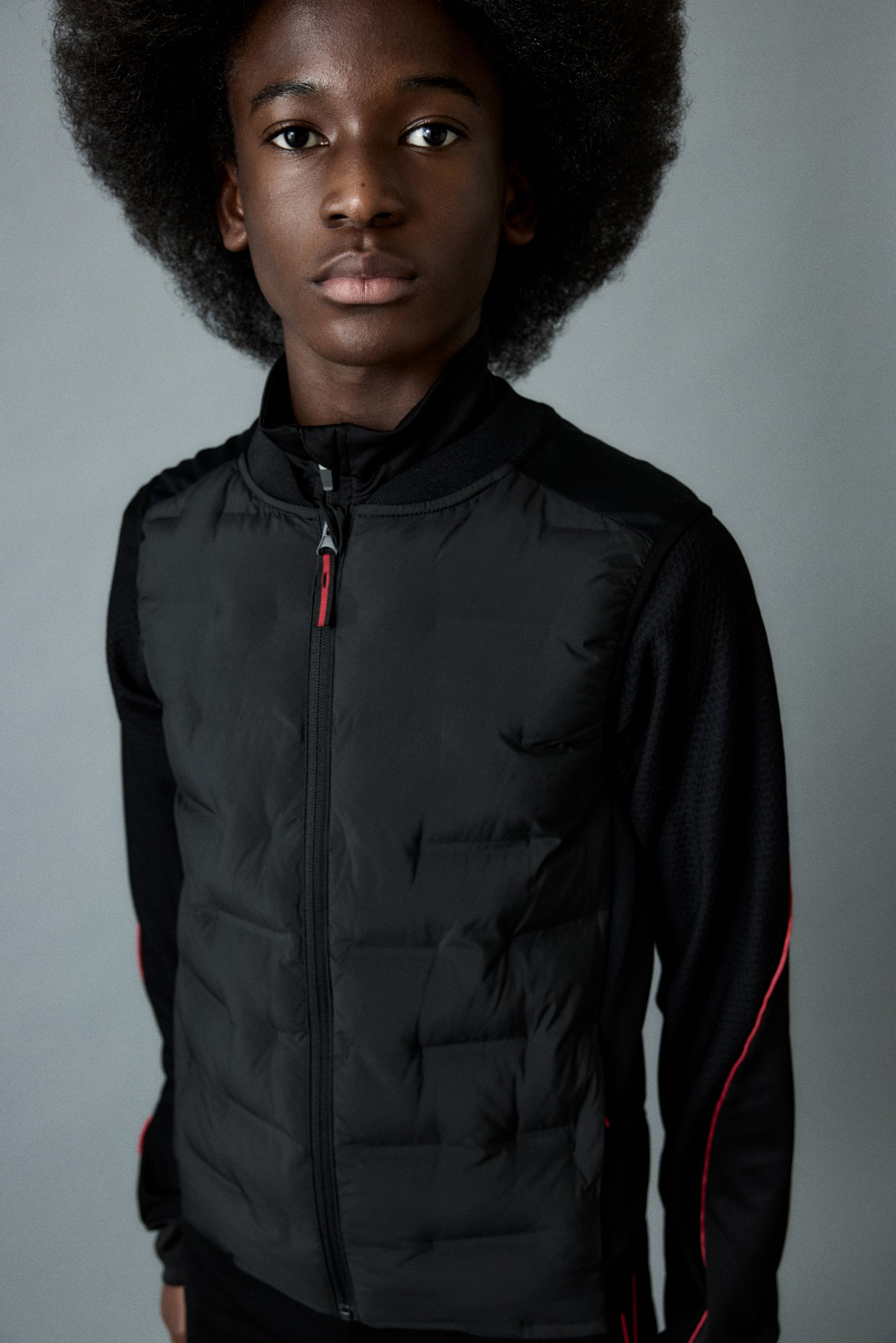 Prikaži veću sliku: Црна јакна има подстavljену предњу плочу, контрастне дуге рукаве од фине мрежасте тканине са црвеним паспулом и ребрасту крагну. Сребрни рајсфершлус са црвеним клизачем затвара јакну.