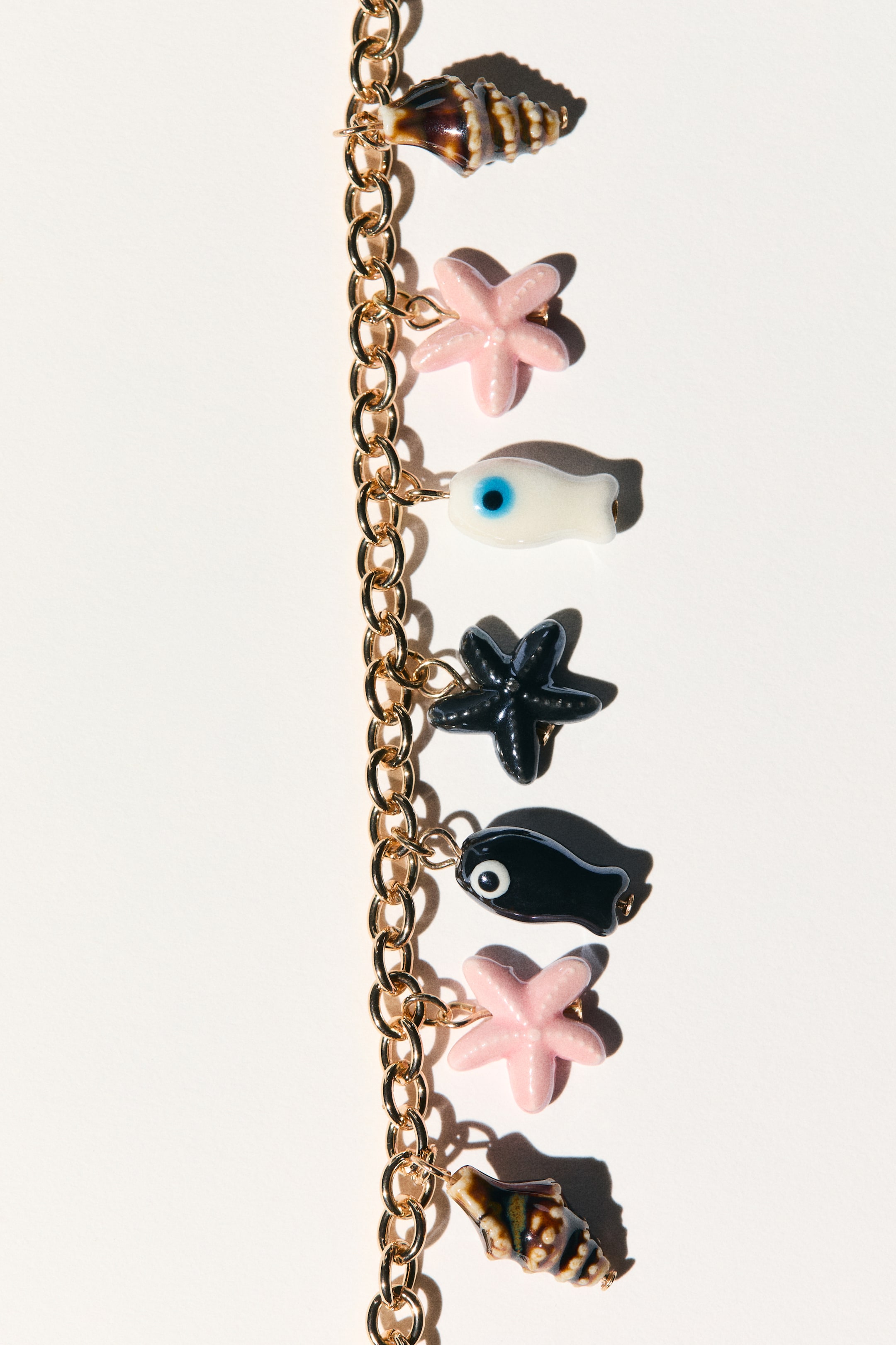 View larger image: A charm bracelet showcasing a gold-toned chunky chain adorned with an array of charms hanging vertically. The charms feature brown and cream striped seashells, pink starfish, a white fish with a blue eye, a dark gray starfish, and a black fish with a white eye.