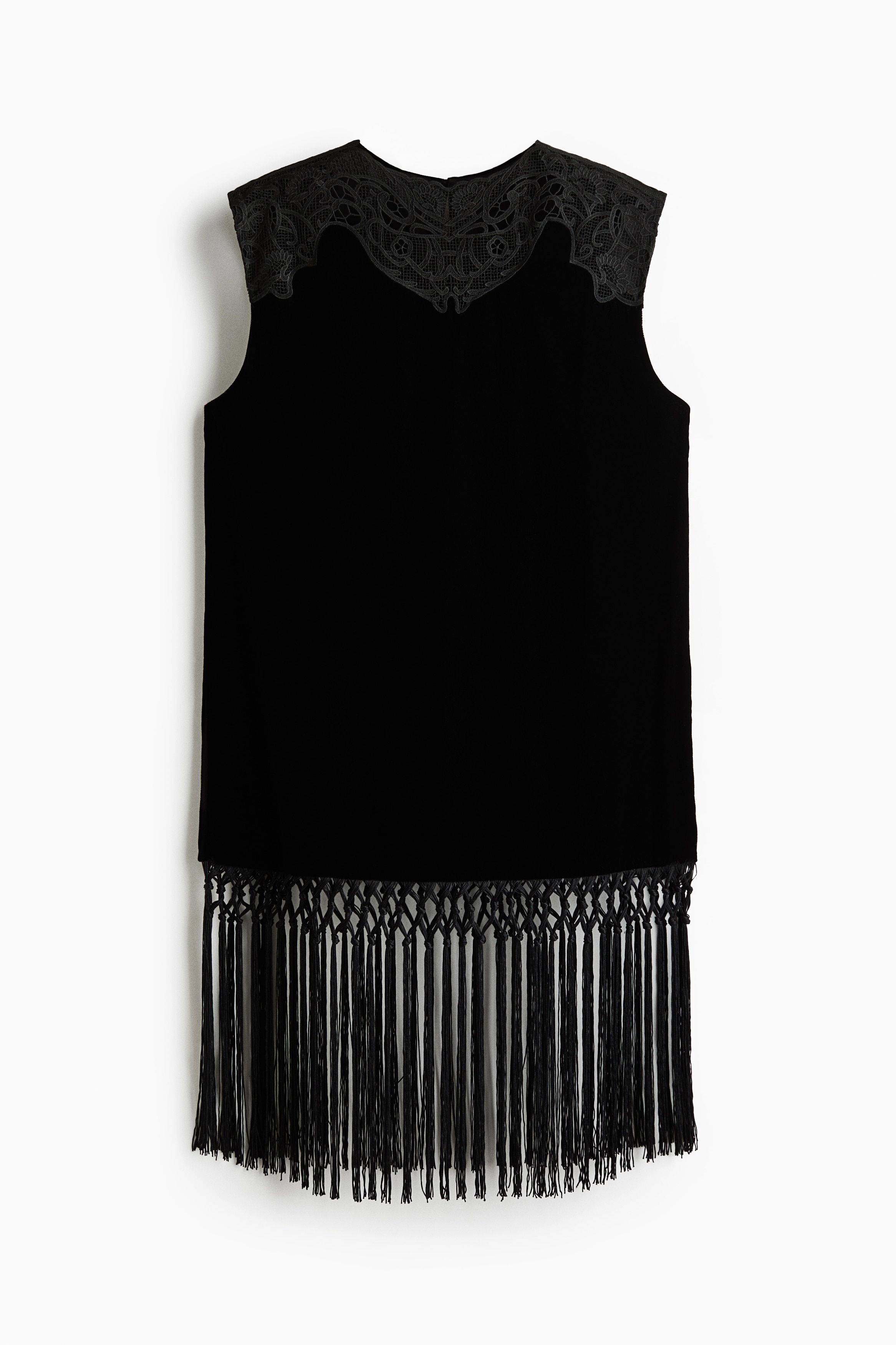 A black velvet sleeveless top featuring a sheer lace yoke at the neckline and a long fringe trim at the hem is being worn by the model. The top is styled with matching black trousers, with the model standing with hands placed in the pockets.