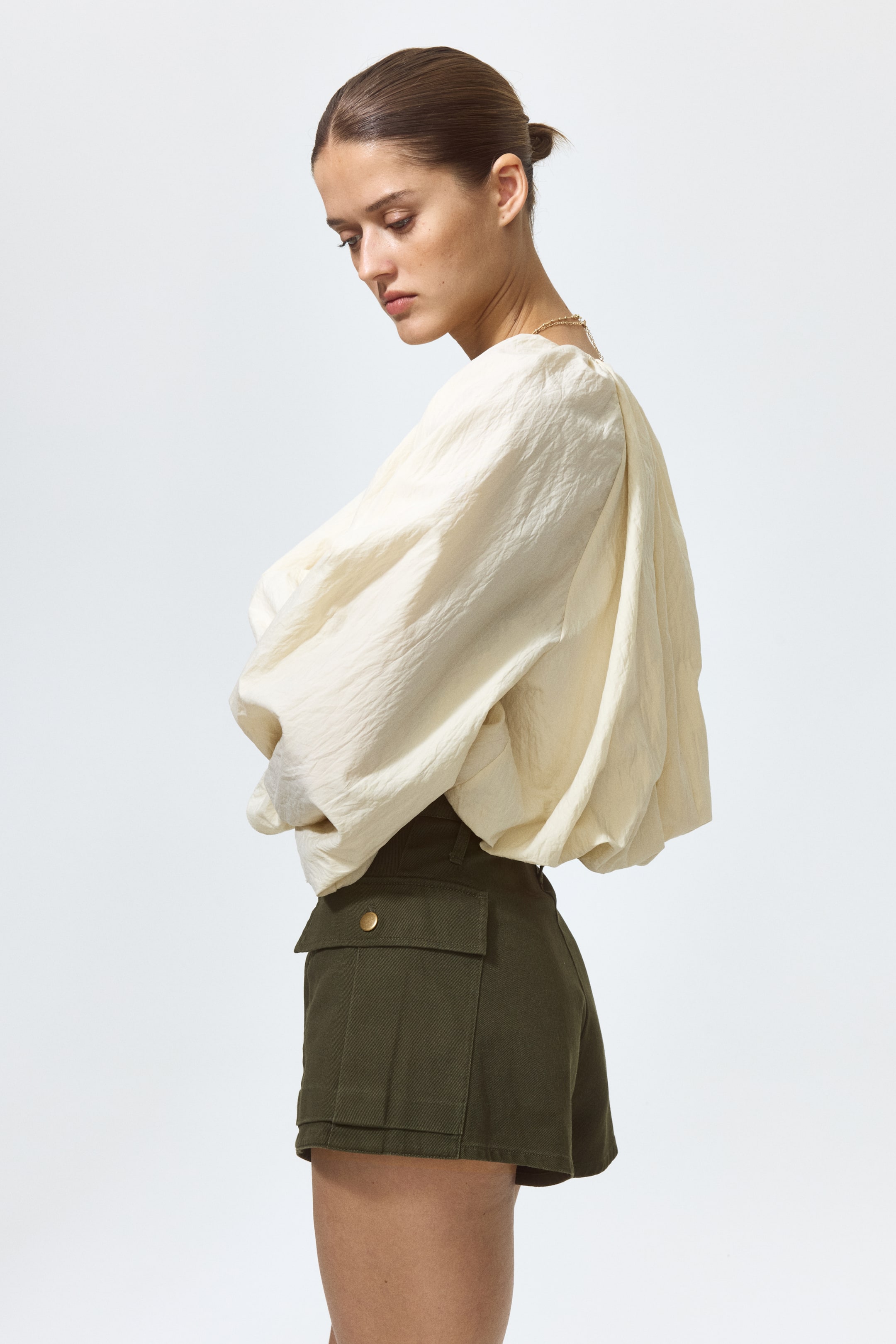 View larger image: The model is wearing olive green cotton twill shorts. They feature a high waist with a button closure and bellow leg pockets with a flap and button.
